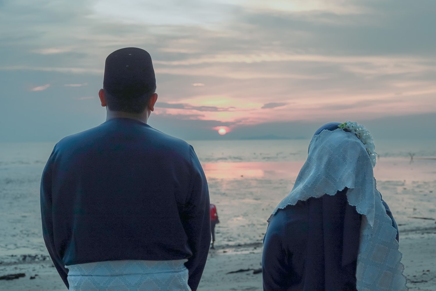 Two people sit together looking out at a peaceful ocean sunset with pink and purple hues in the cloudy sky.