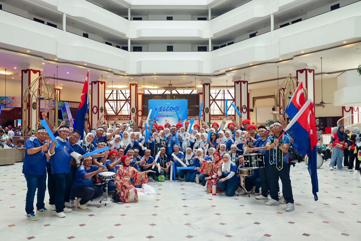 Large group in blue and white uniforms with flags posing in hotel atrium at corporate event.