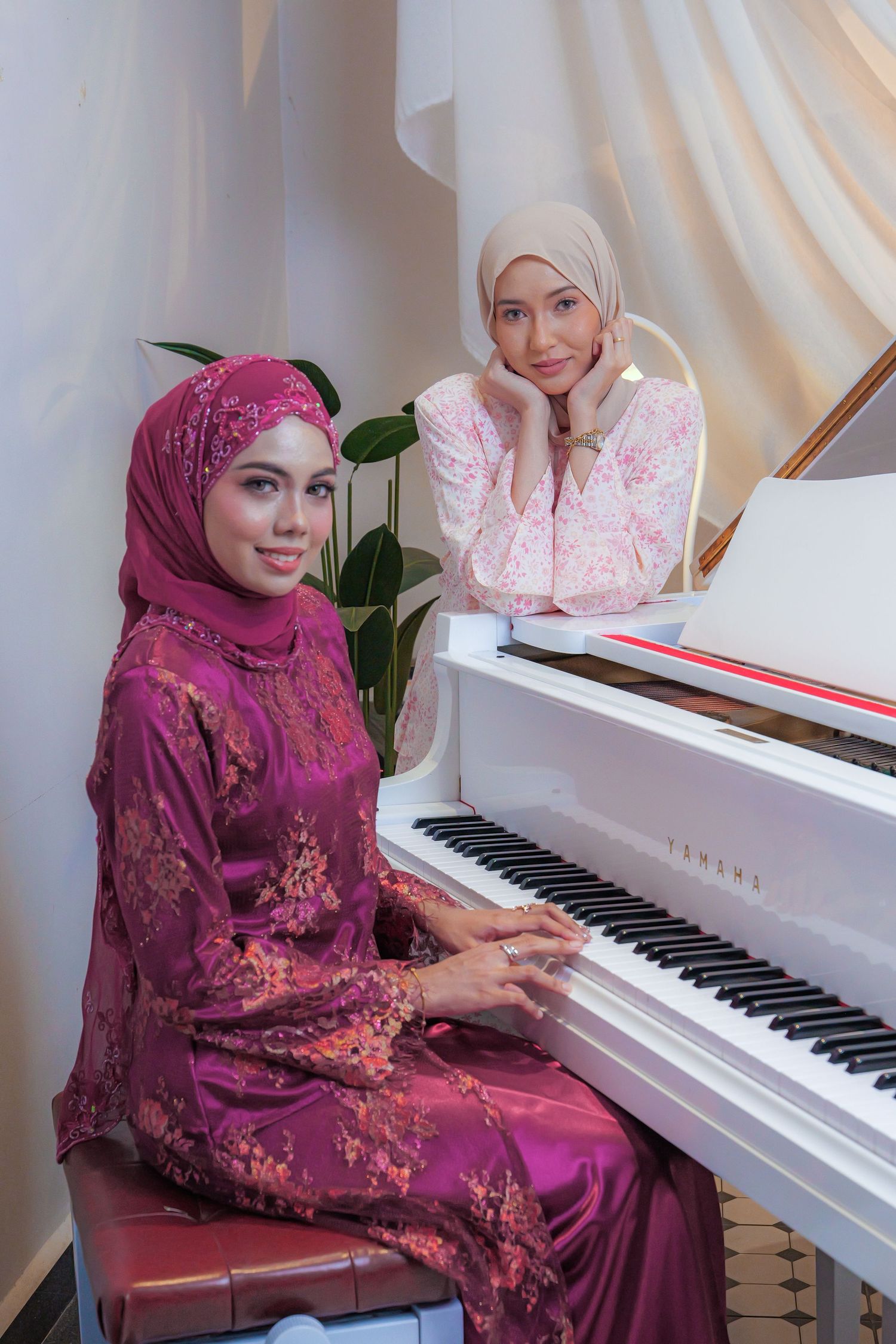 Two women in hijabs sitting at a white piano, one playing while wearing a burgundy floral dress and the other observing.