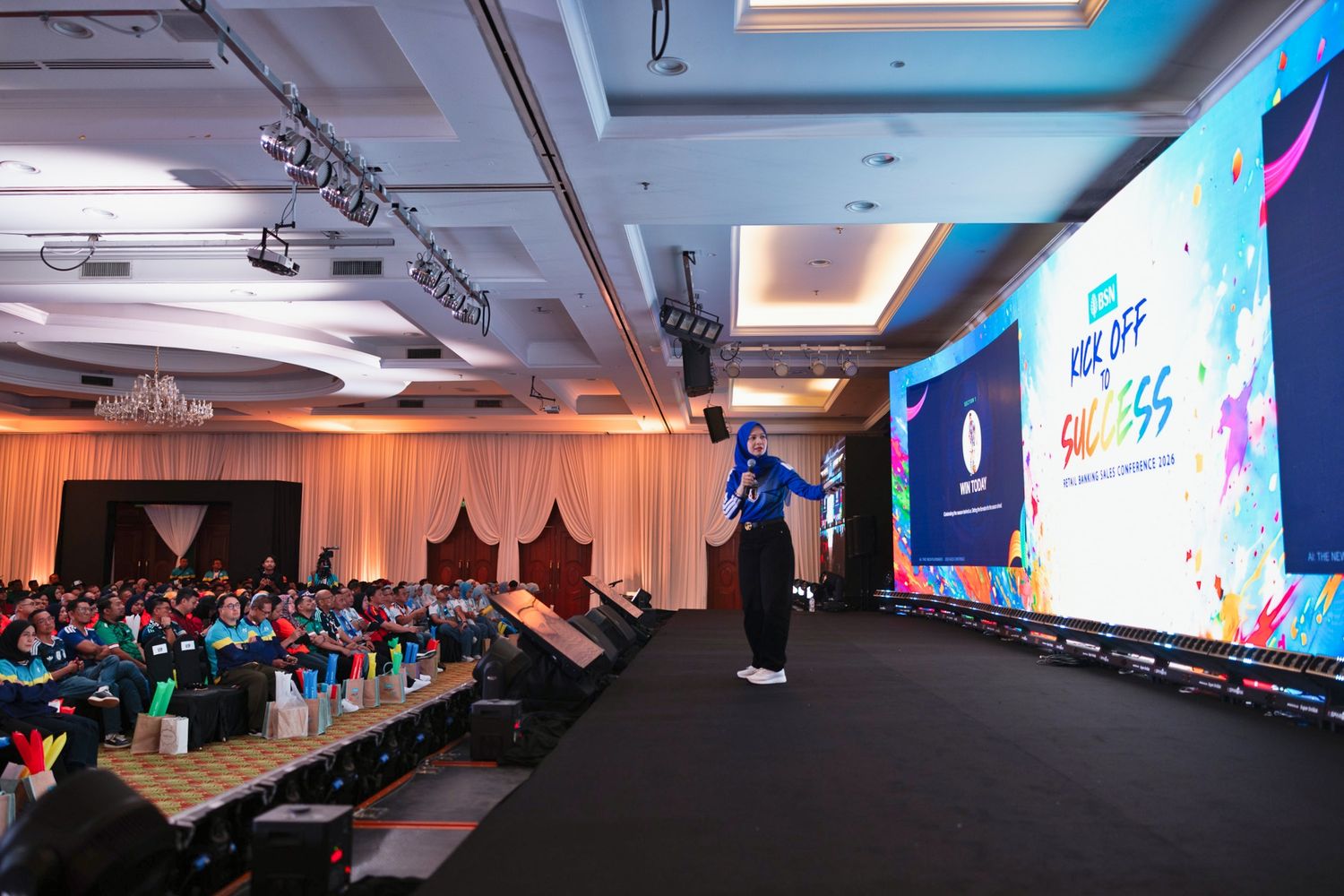 Speaker presenting on stage at a colorful corporate conference with large LED screen and seated audience in ballroom.