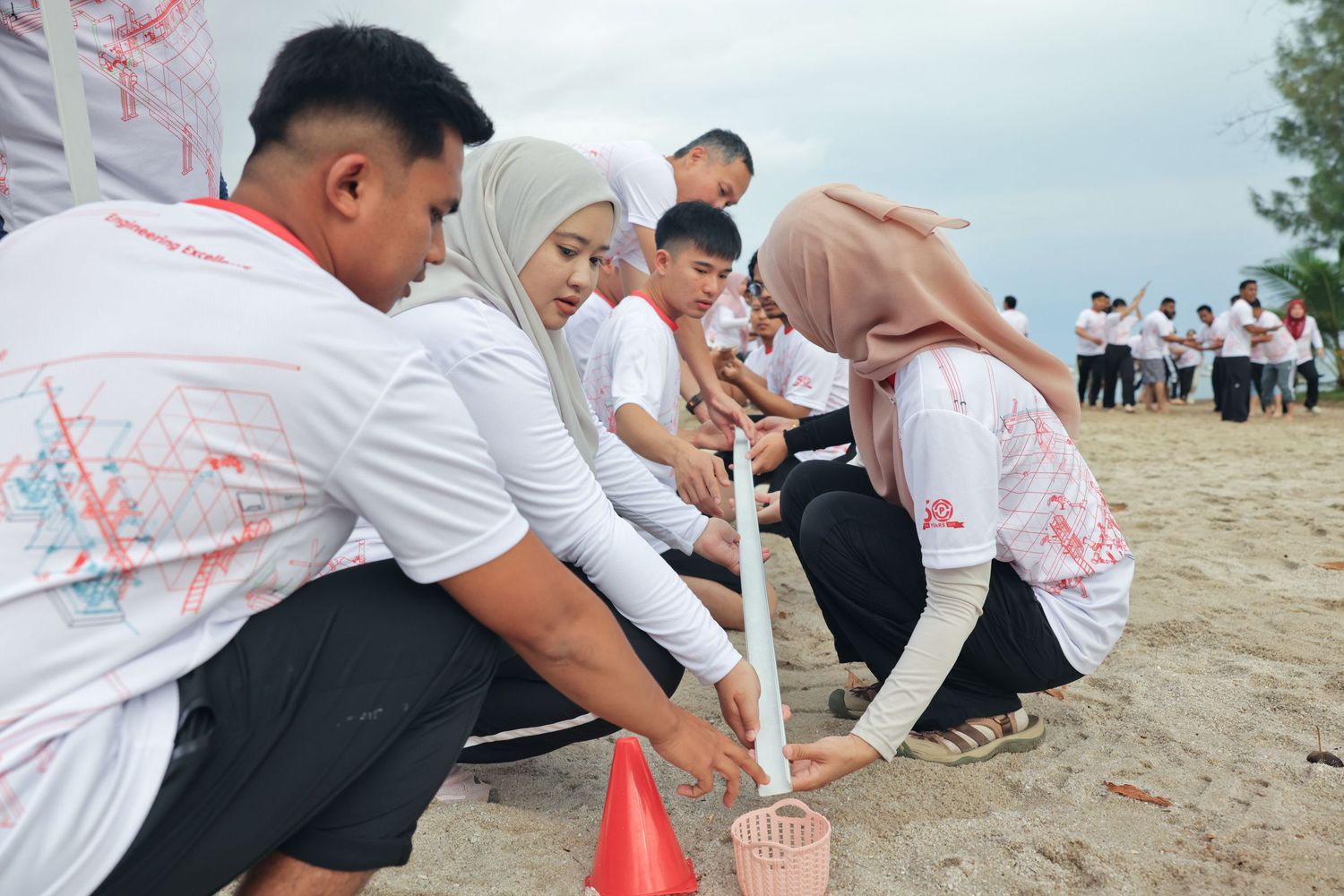 Group of people in white shirts participating in a beach team building activity, digging in sand near a red cone marker.