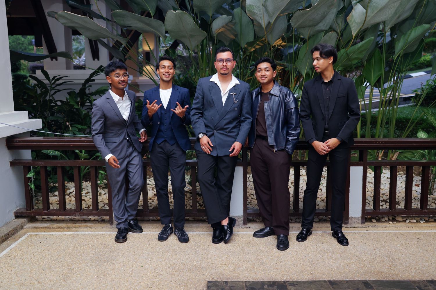 Five well-dressed young men posing together in suits outdoors near tropical garden greenery.