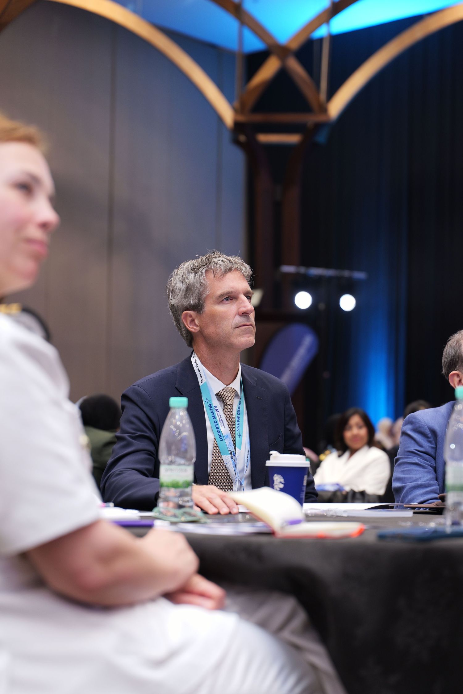 Business conference attendee in navy suit sits attentively at table with water bottle during professional event.