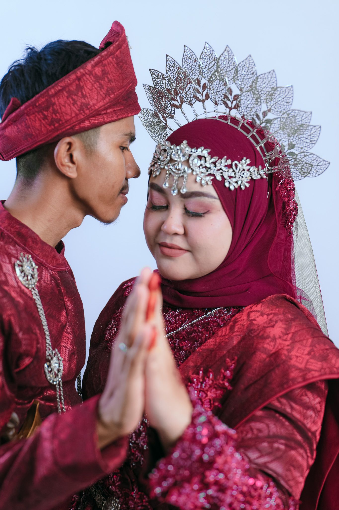 A couple in traditional maroon wedding attire with a bride wearing a decorative hijab and matching crown headpiece.