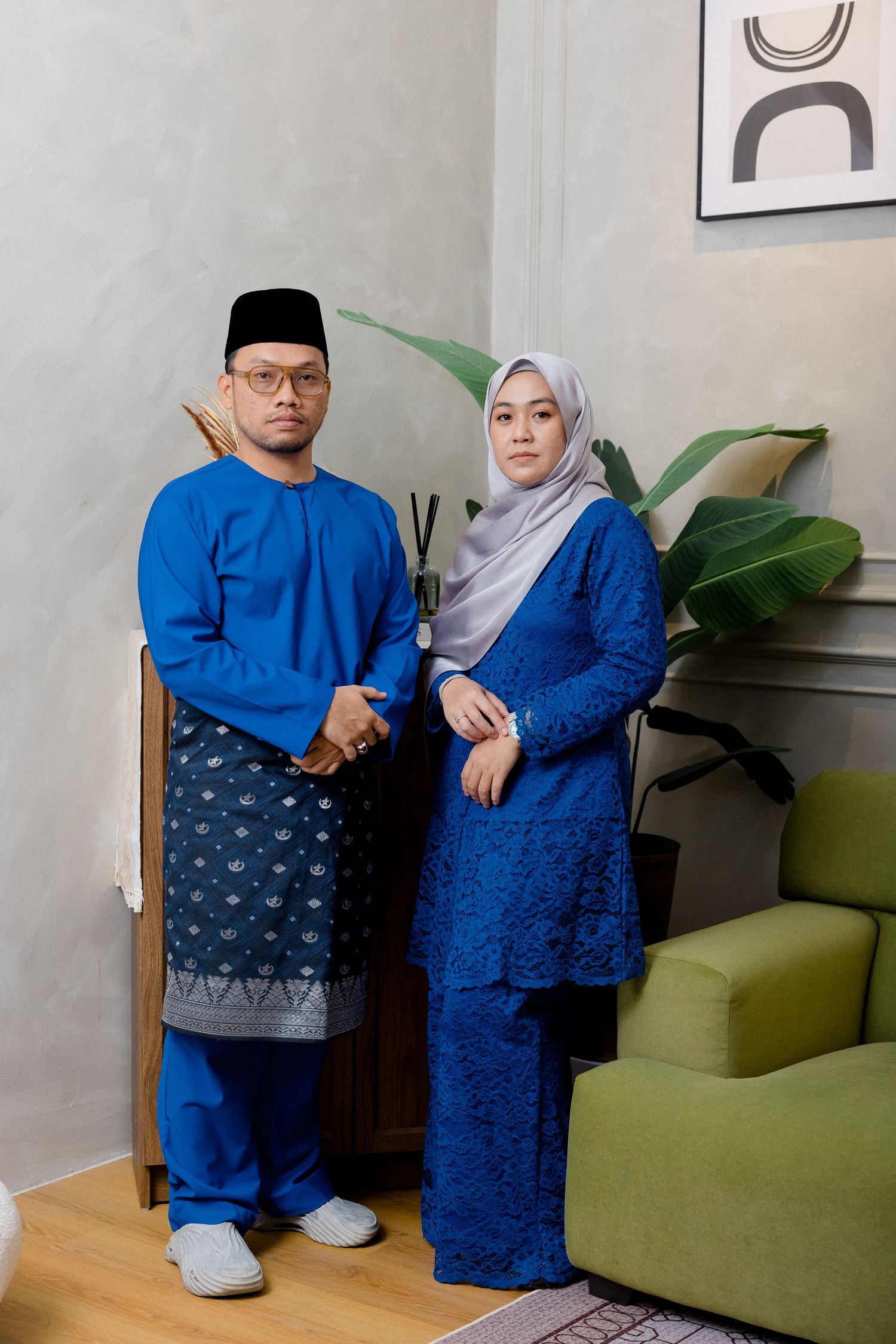 A couple wearing coordinating blue traditional outfits stands together in a living room with a green couch.