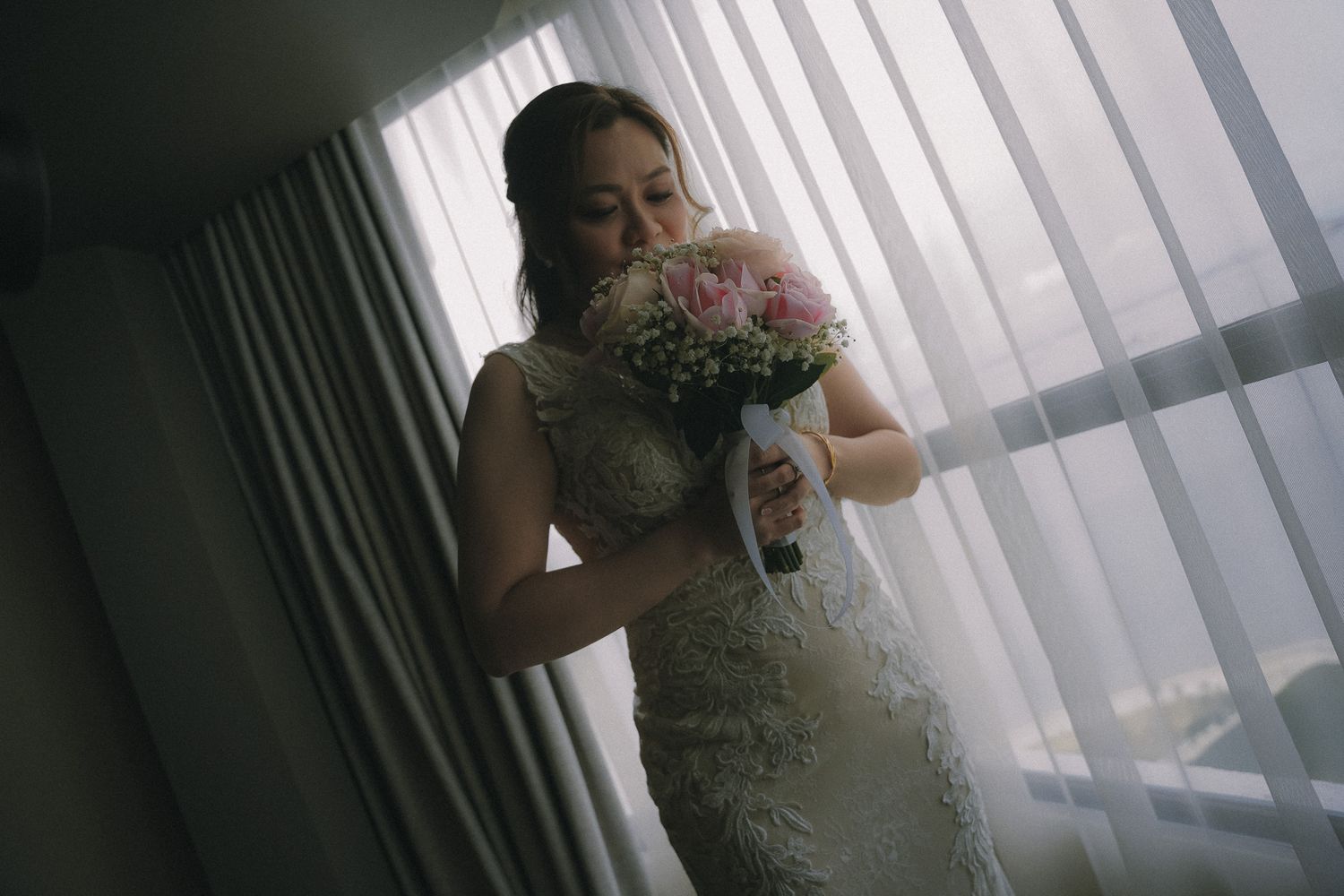 A silhouetted figure in a lace wedding dress stands by a window holding a pink bouquet in moody lighting.