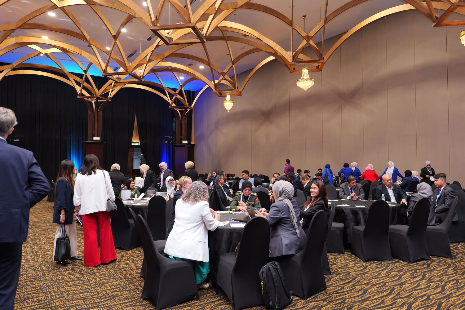 Conference attendees seated at round tables in an elegant ballroom with wooden beam ceiling and pendant lighting.