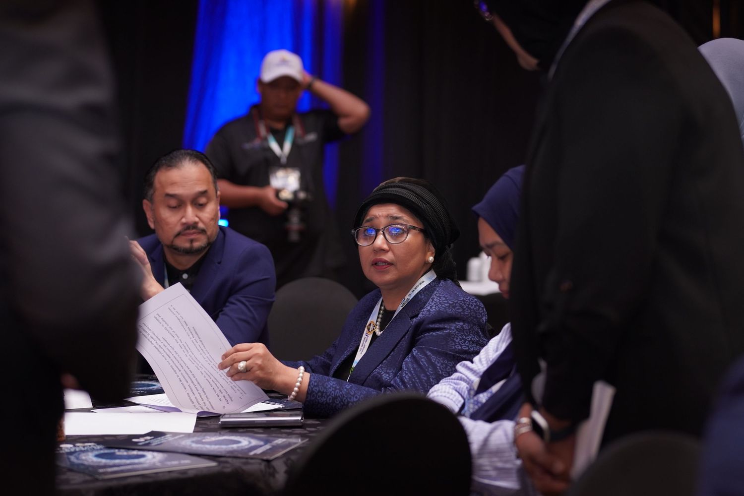 People seated at tables review papers during what appears to be a professional meeting or conference event with blue lighting.