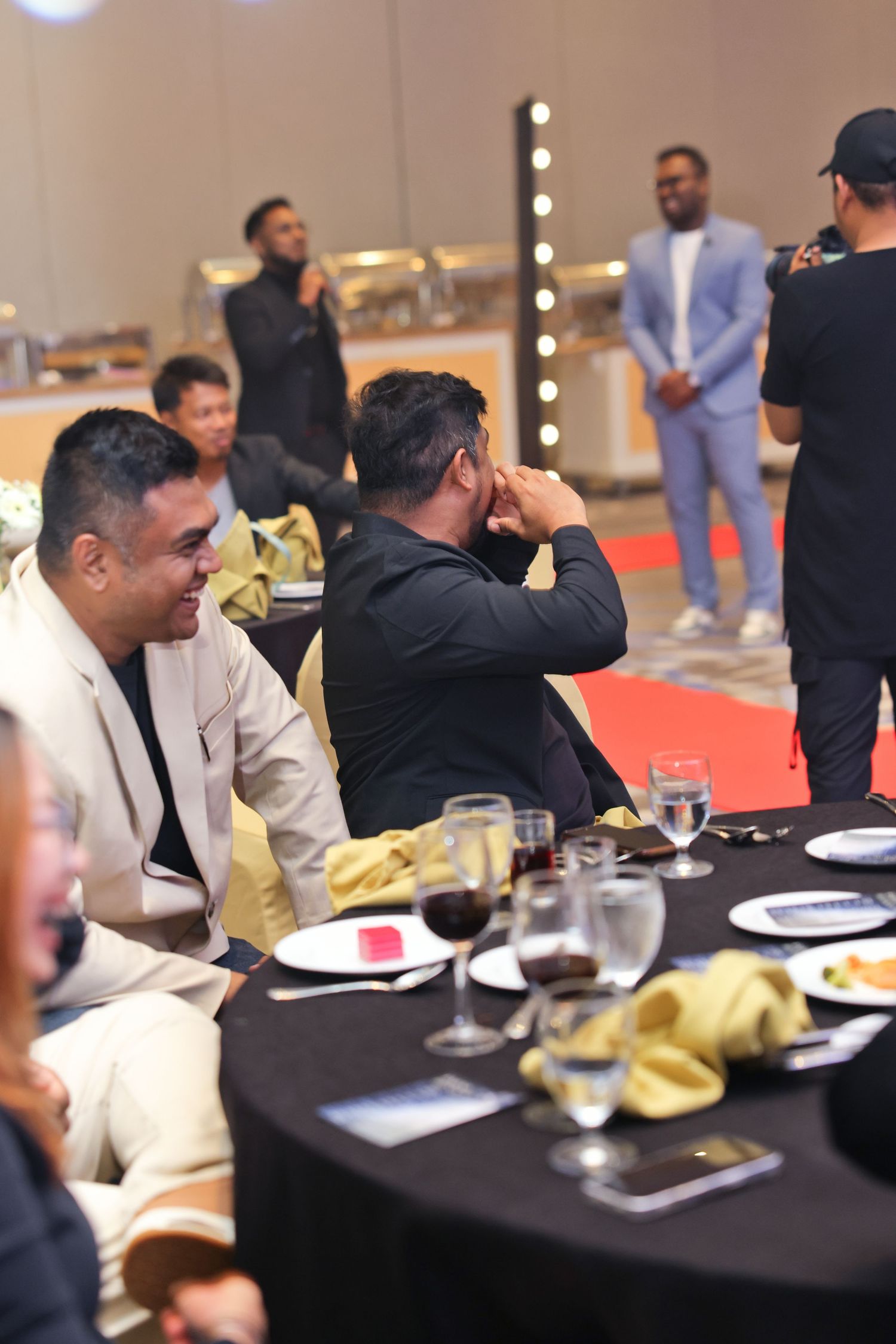 Guests seated at a formal banquet table with wine glasses, while others stand on a red carpet at an elegant event.