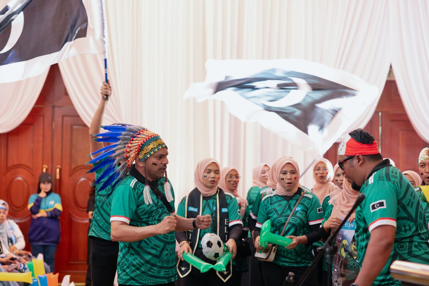 People in green jerseys wave flags and hold a soccer ball at a festive indoor sports celebration event.