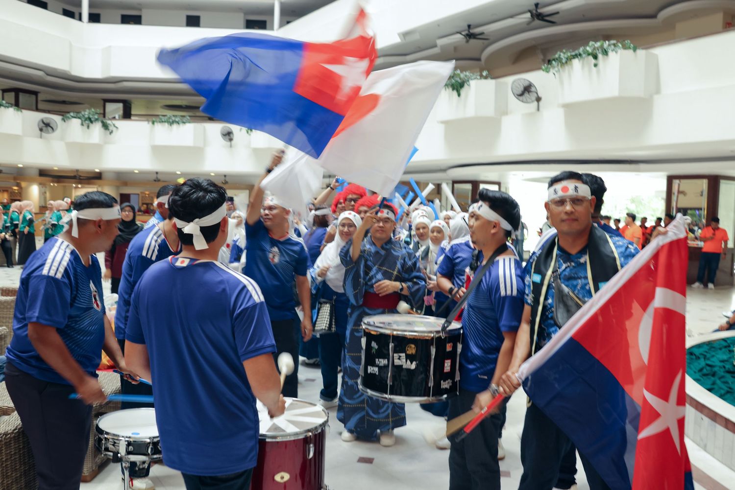 Energetic sports fans in blue uniforms wave flags and play drums in a festive indoor rally celebration.