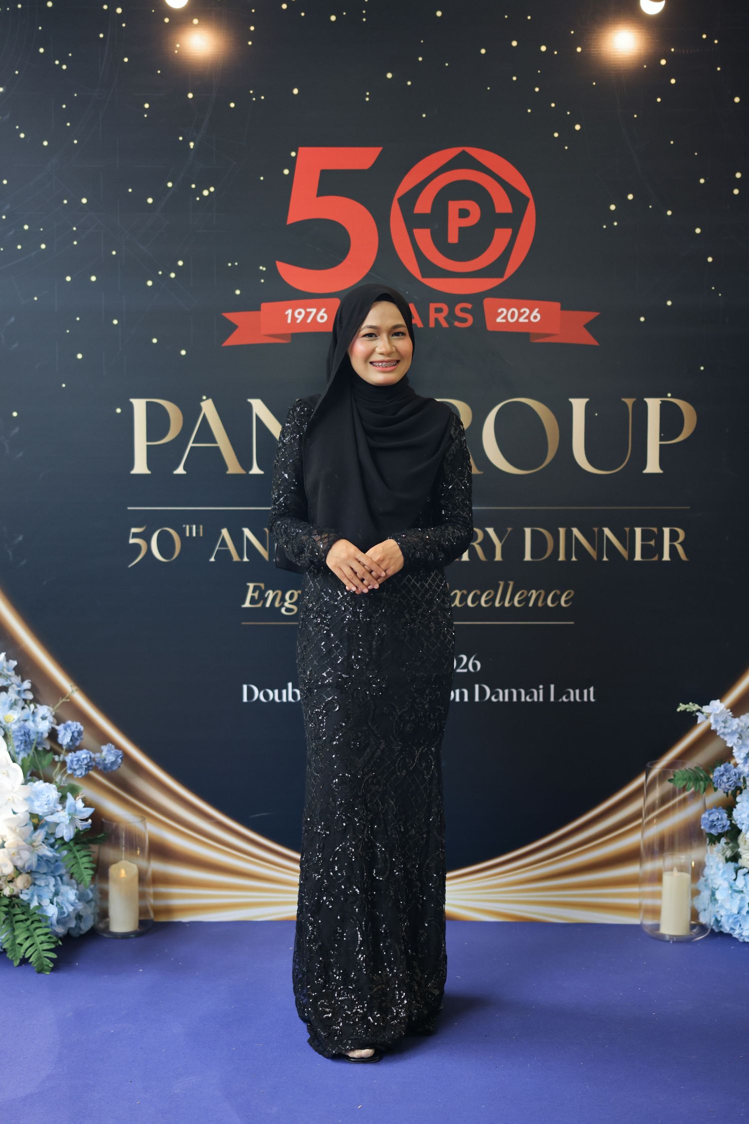 Woman in black hijab and sequined gown poses at Pana Group 50th Anniversary Dinner backdrop.