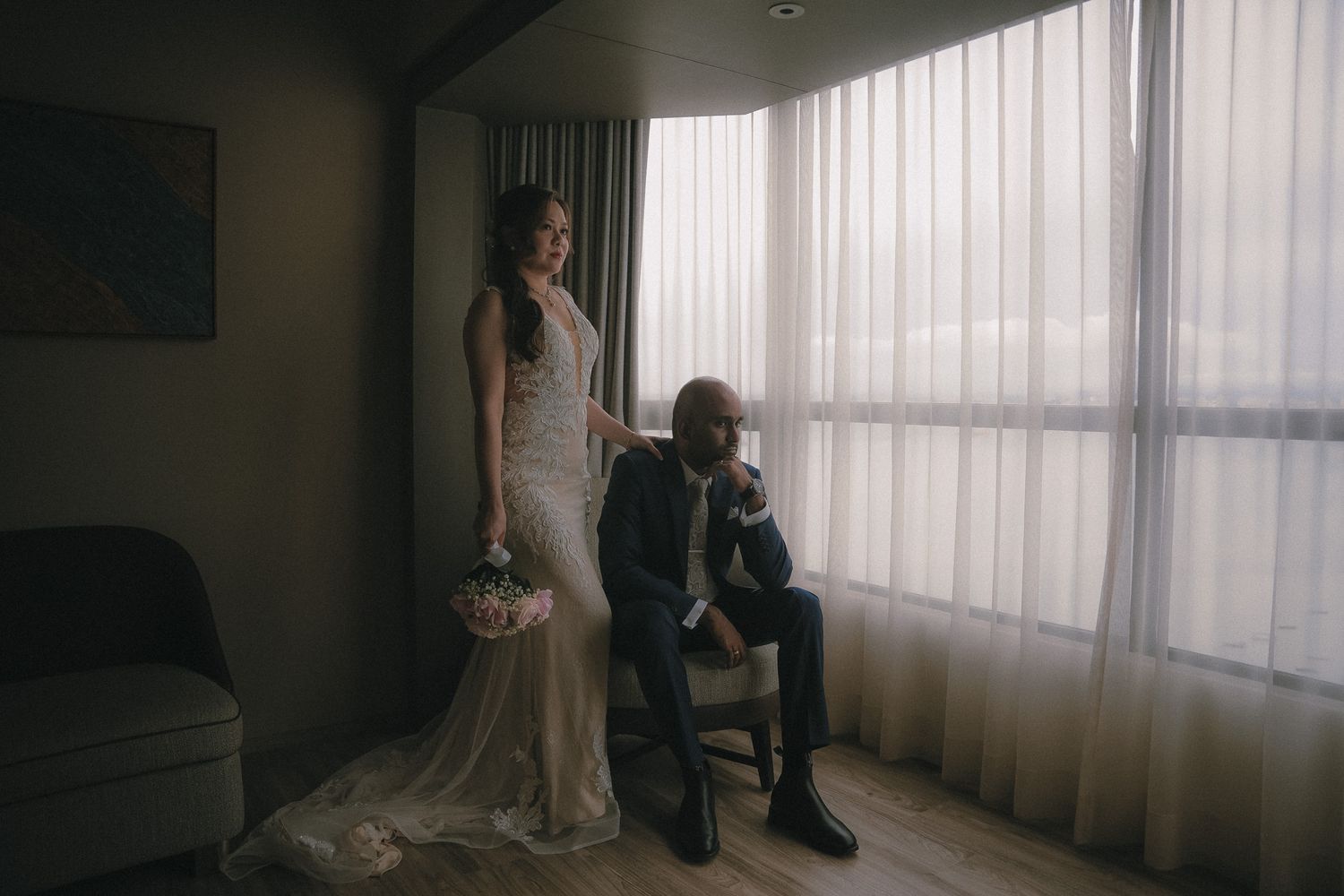 Silhouetted couple in elegant wedding attire poses by a bright window in a contemporary room.