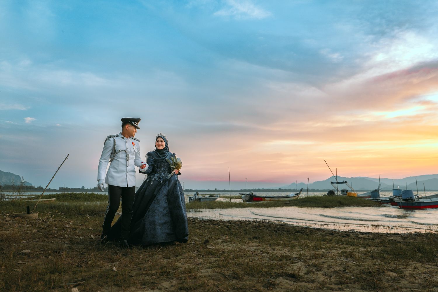 A couple in vintage period attire stands together on a beach at sunset with boats in the background.