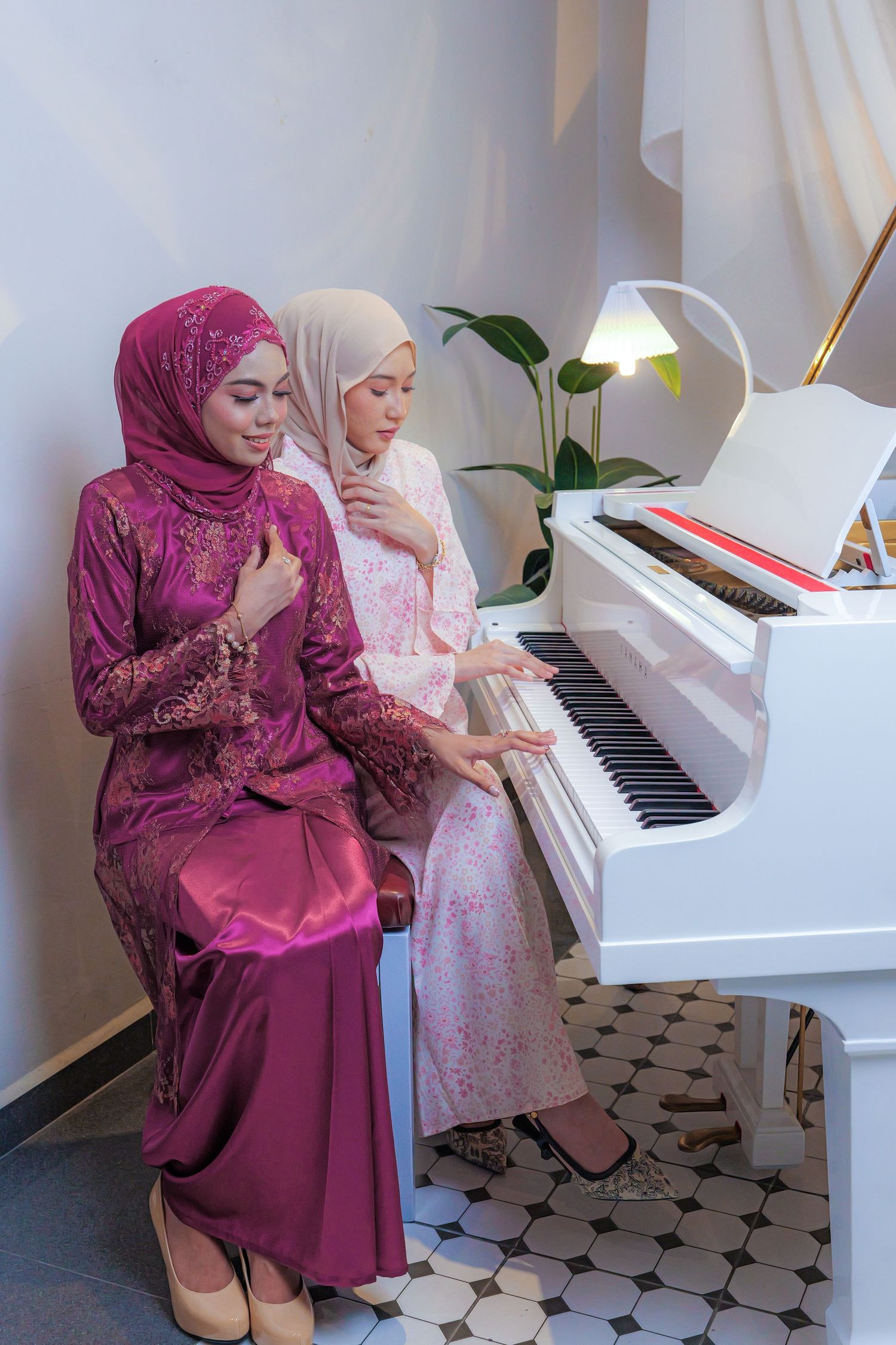 Two people in formal attire pose together at a white piano against white draped backdrop.