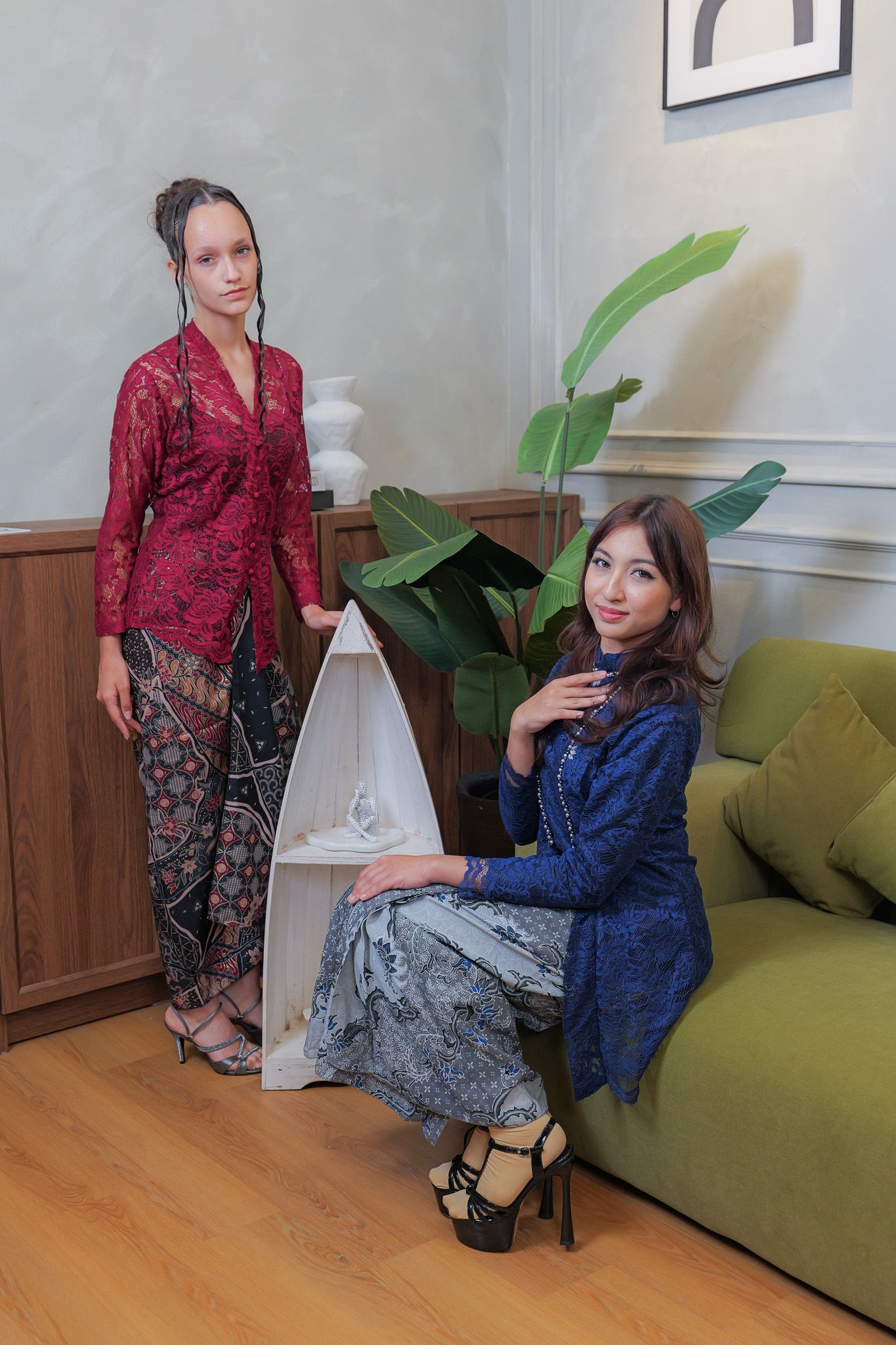 Two women in contrasting outfits pose in a modern living room with a green couch and potted plant.