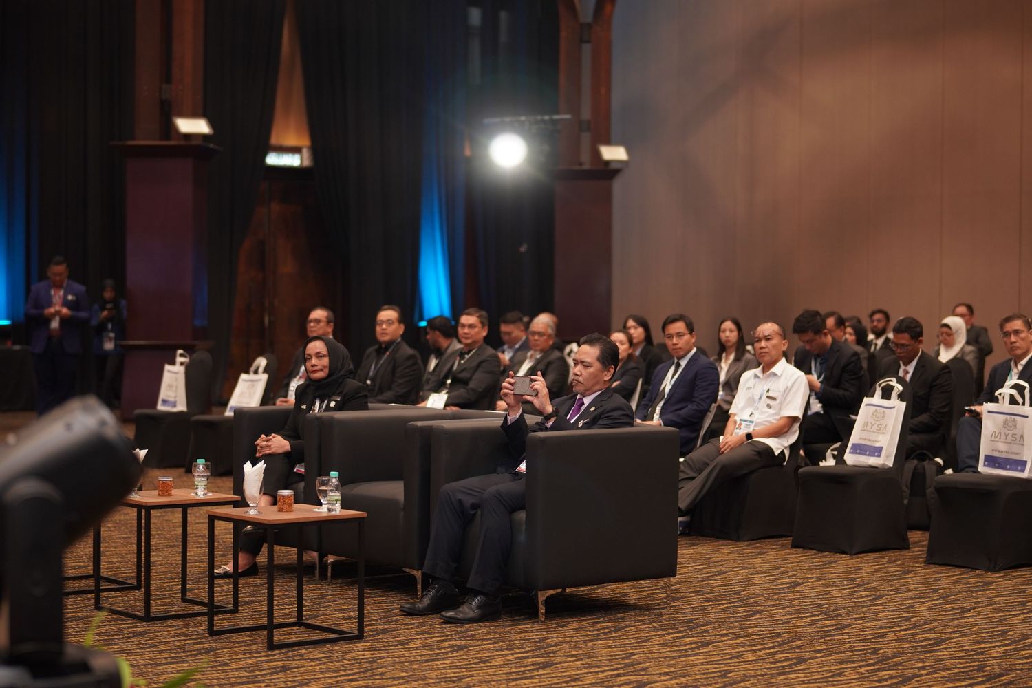 Wide shot of a formal business conference hall with attendees seated in rows facing the stage.