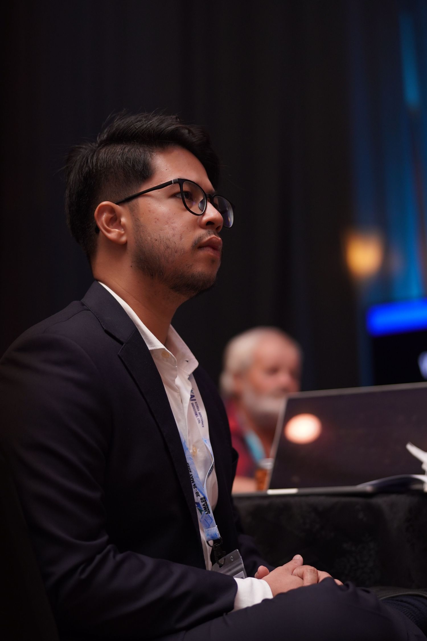 Attendee in business attire and glasses sits attentively during conference presentation.