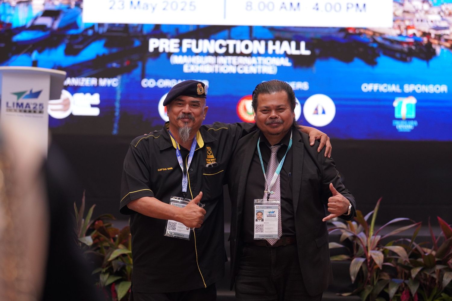 Two attendees in black attire pose together at pre-function hall during conference.