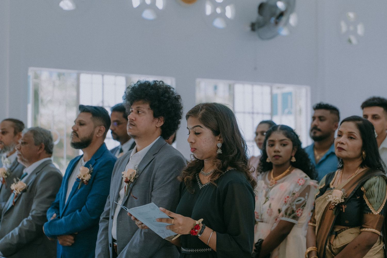 Wedding guests stand together during ceremony while holding programs in an indoor venue with floral decorations.
