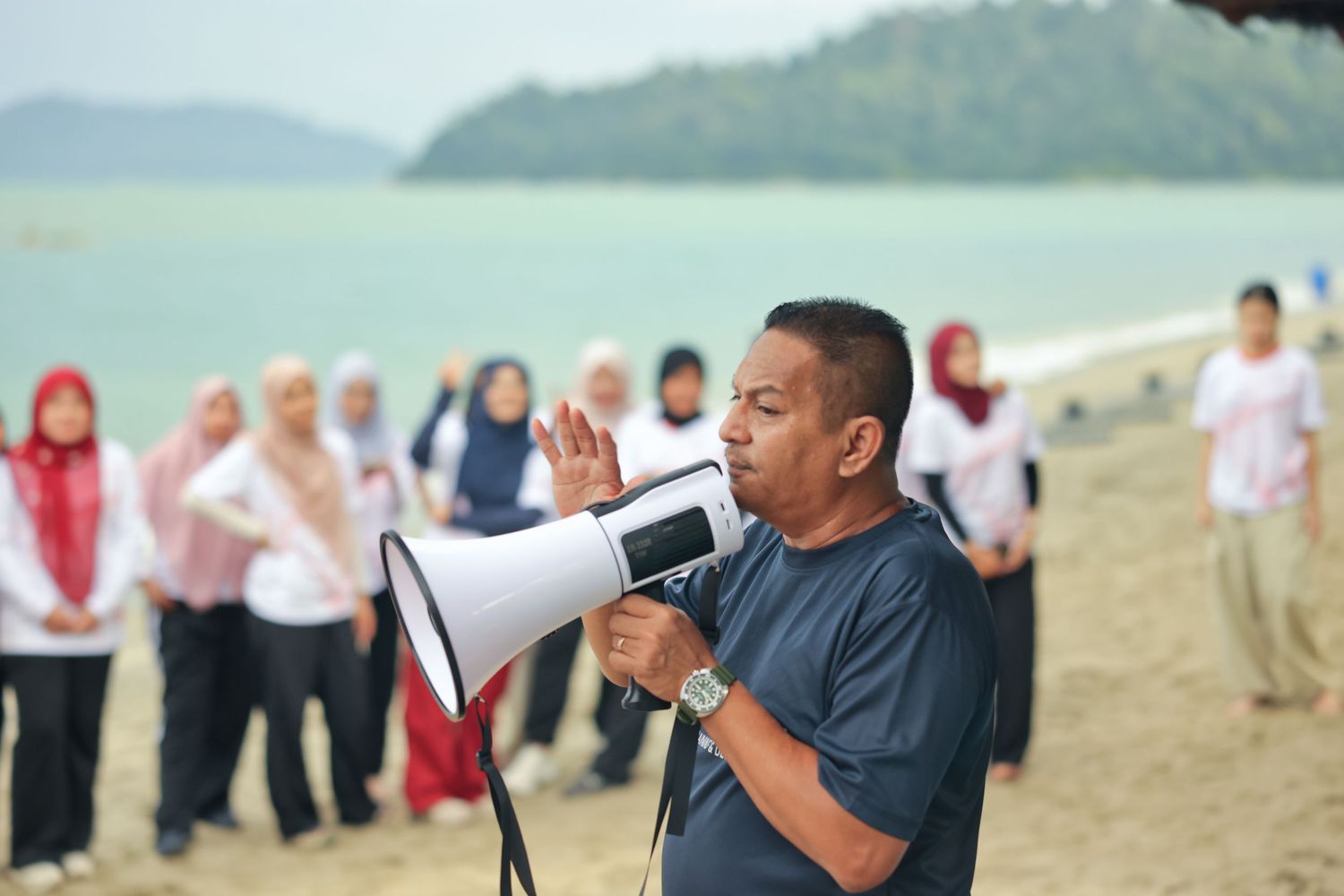 A man speaks into a megaphone on a beach while a group of people in hijabs listen attentively behind him.
