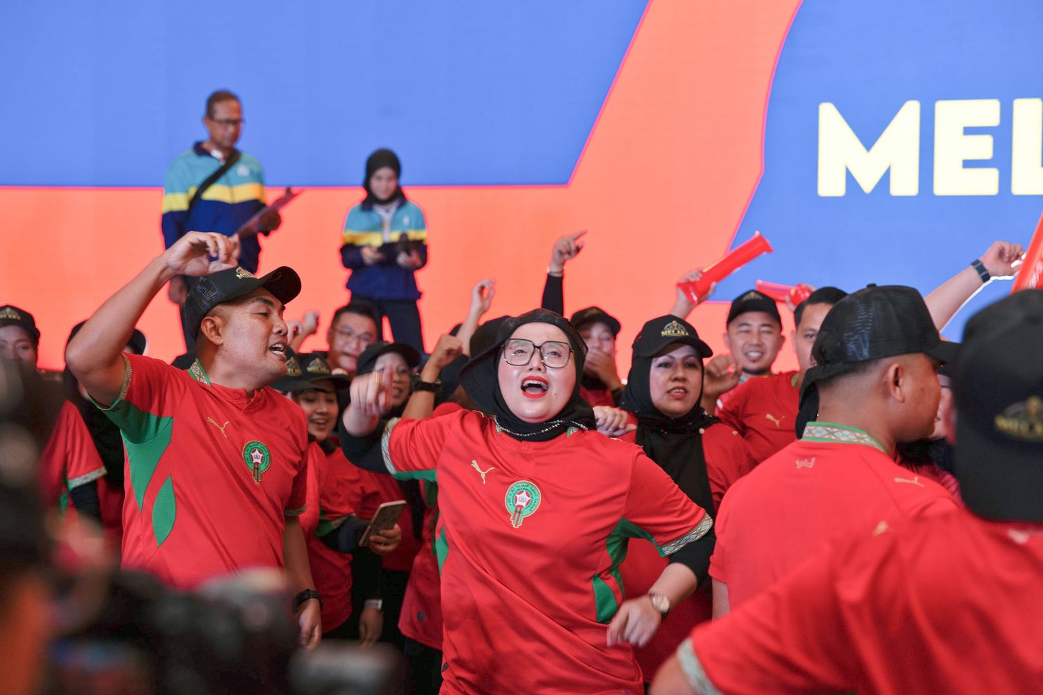 Enthusiastic fans in red Morocco jerseys cheering at a lively outdoor sports event.