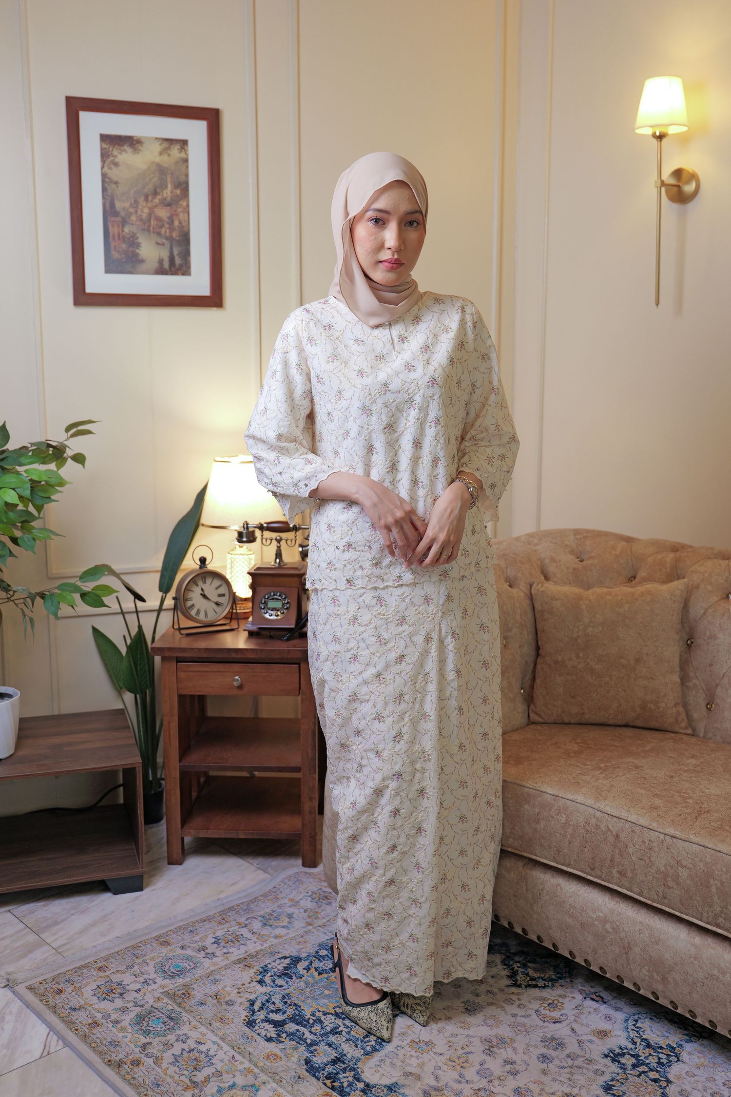 Person in a white floral dress stands on a patterned rug in a cozy living room with warm lighting and houseplants.