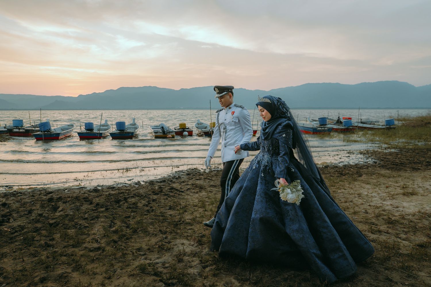 Couple in formal attire walking along beach with boats and mountains in background at sunset.