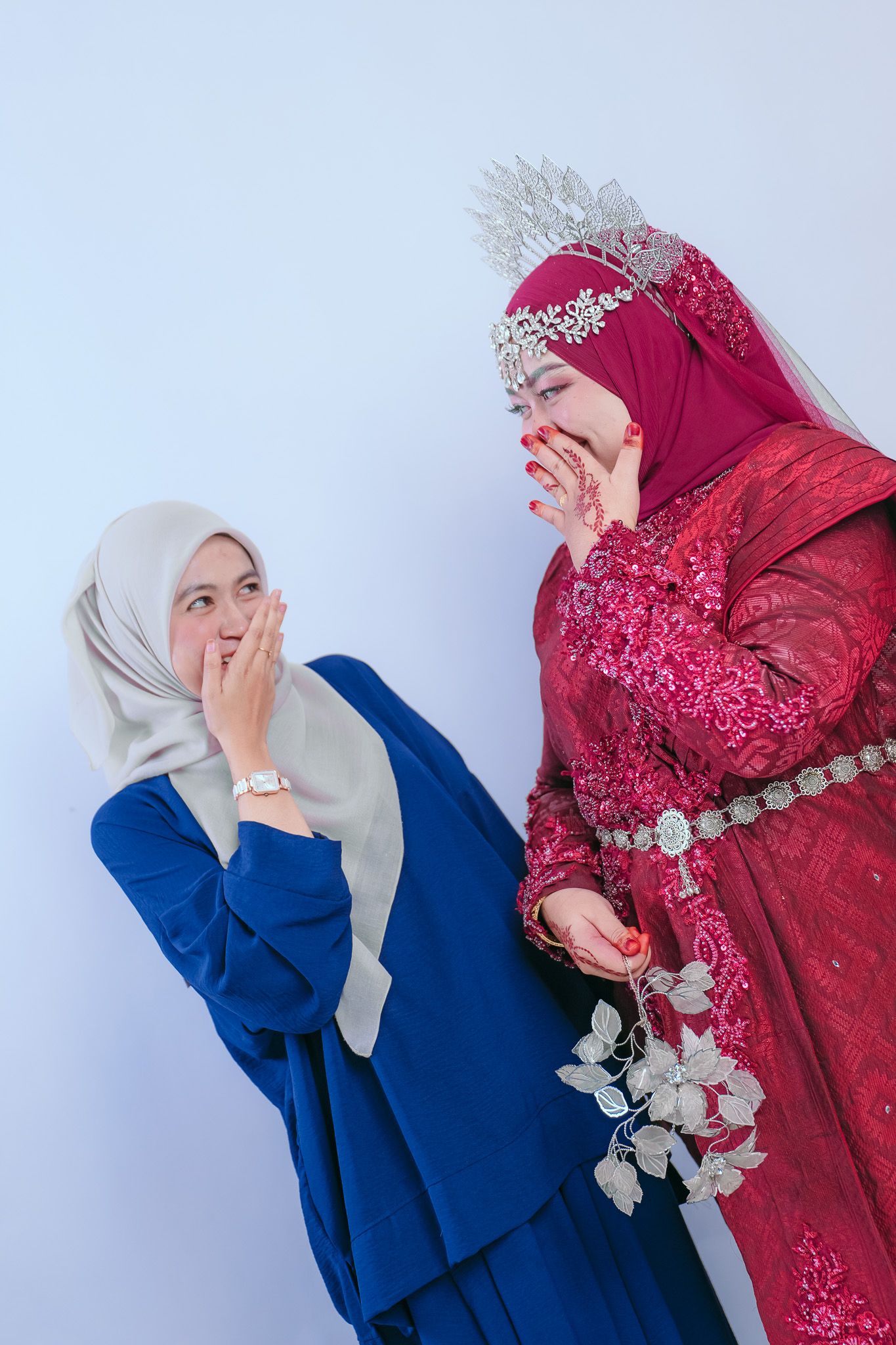 Two figures in traditional modest dress share a joyful moment against a white background, wearing hijabs in blue and red.