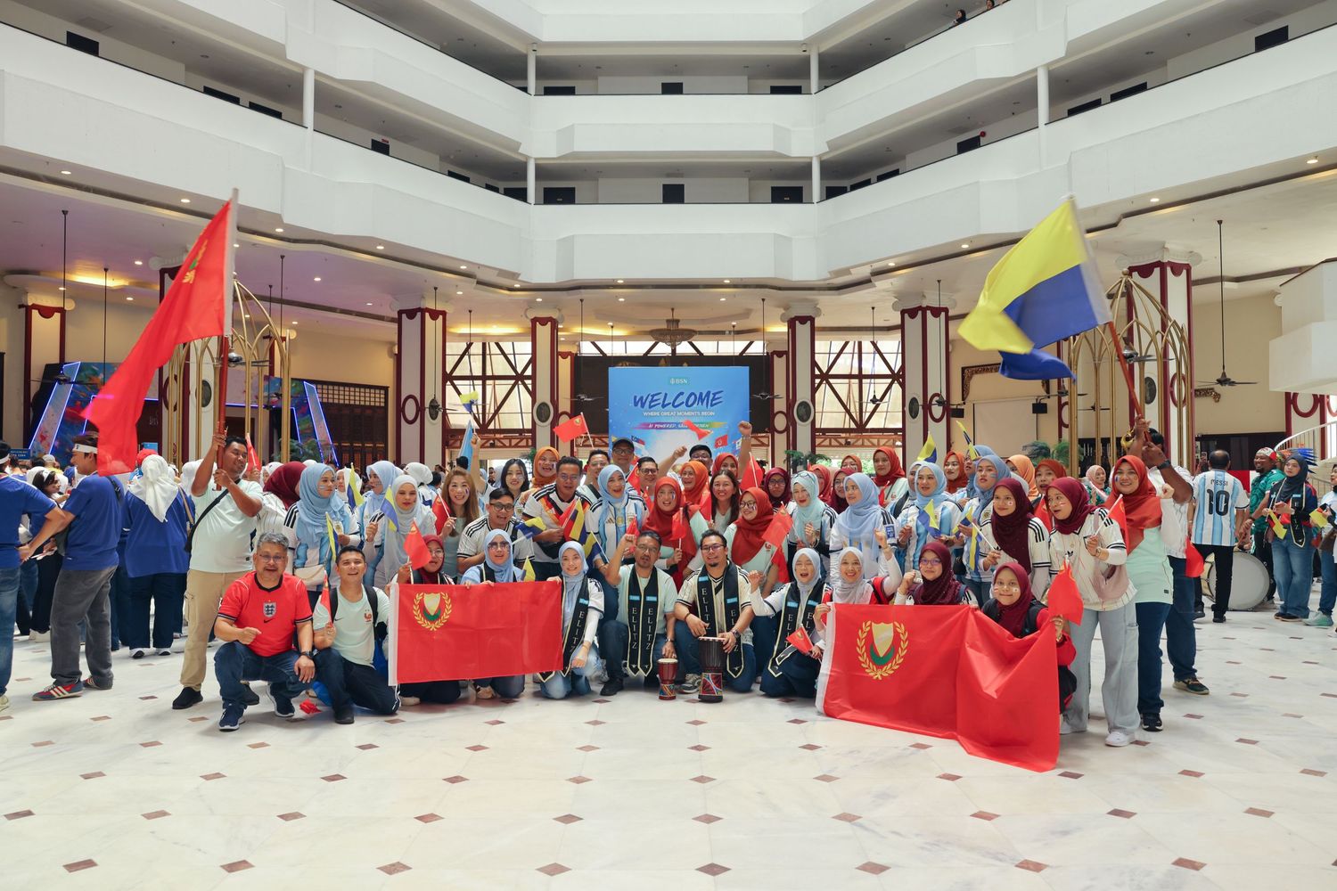 Large group of people posing with colorful flags in a hotel lobby under a Welcome sign.