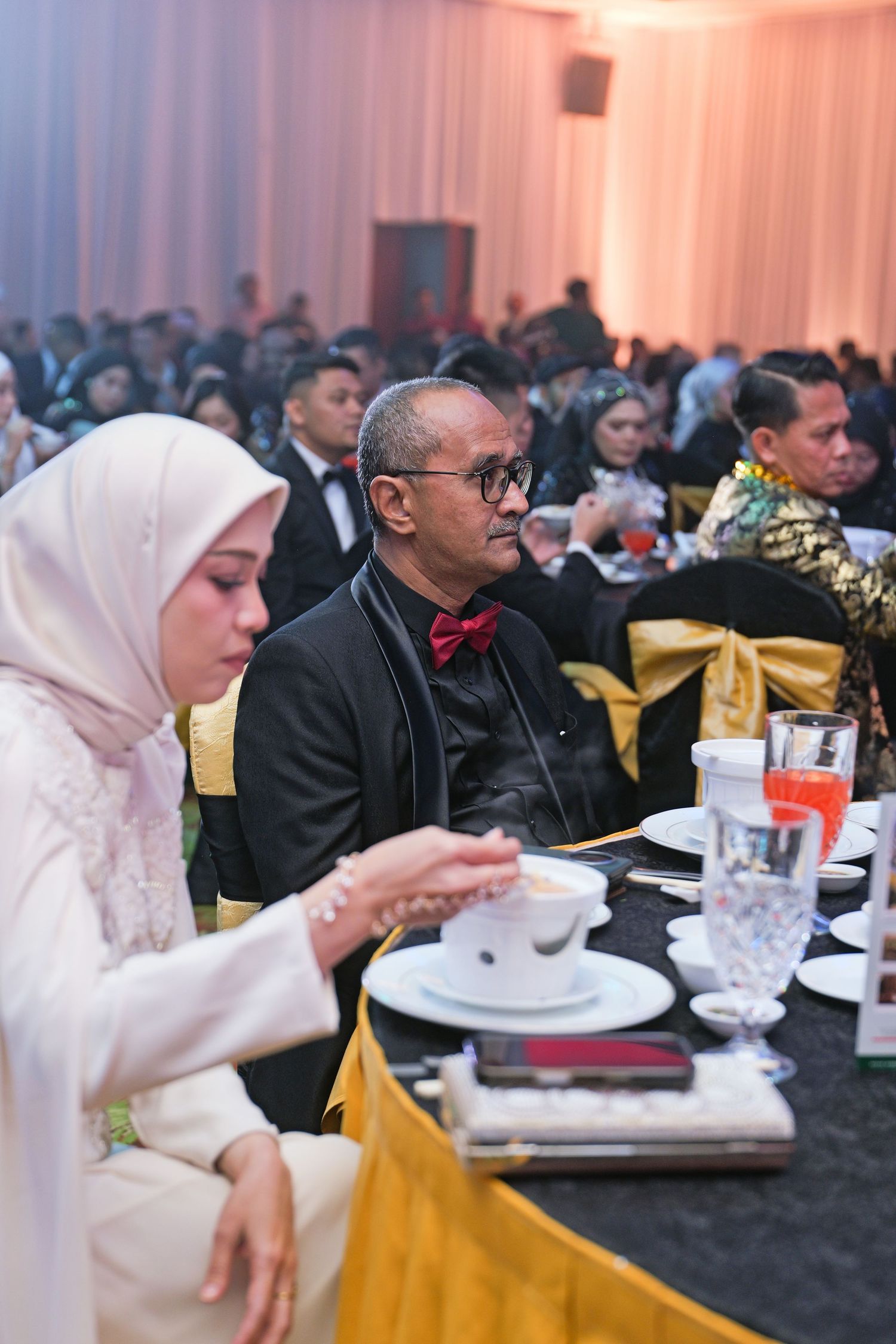 Guests in formal attire seated at a gala dinner table, woman in hijab holding a warm beverage.