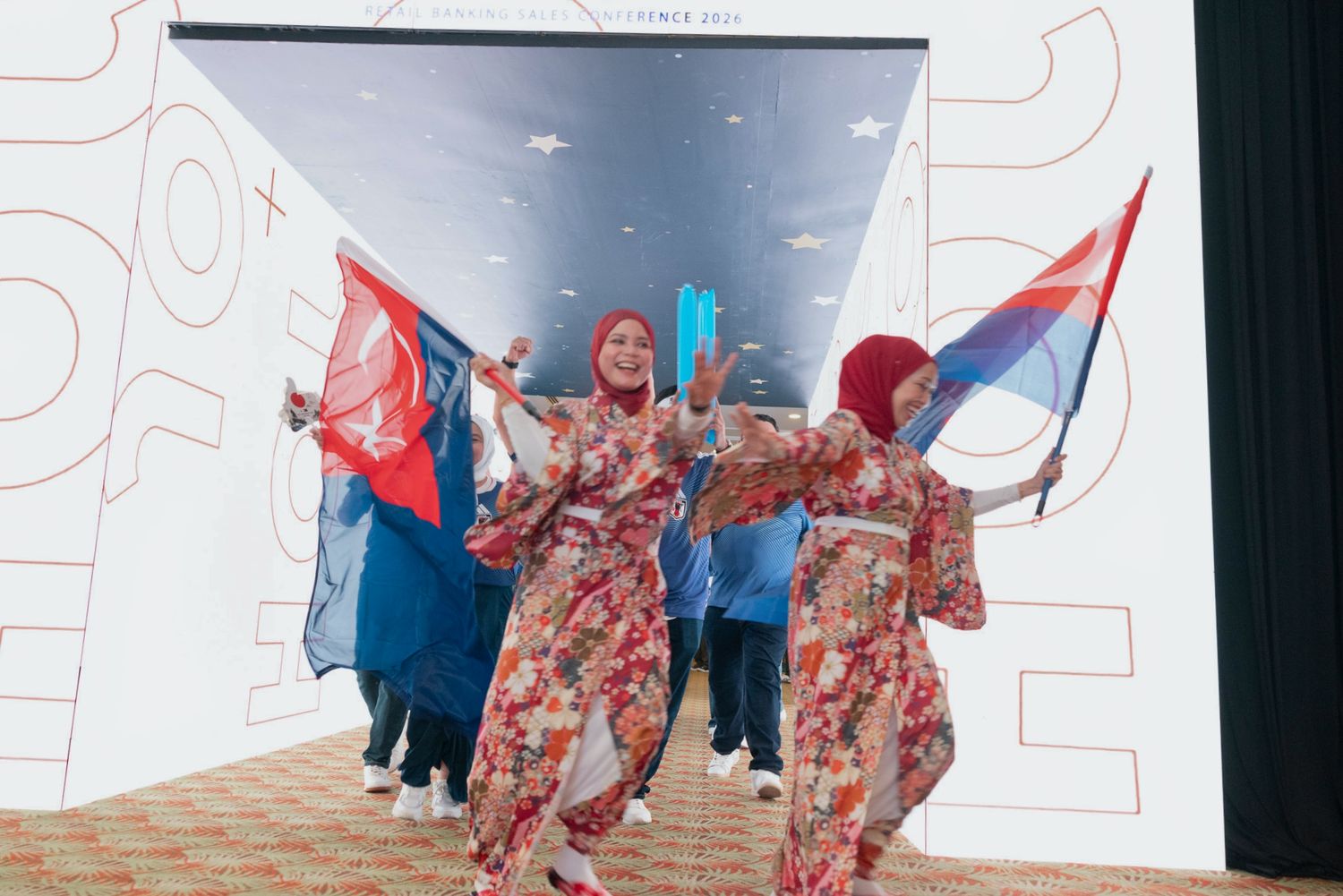 Women in colorful floral outfits wave red and blue flags during a festive cultural performance on stage.