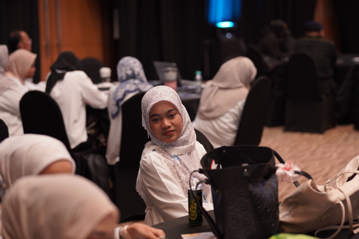 Group of people in white hijabs and clothing seated at tables during an indoor gathering event.