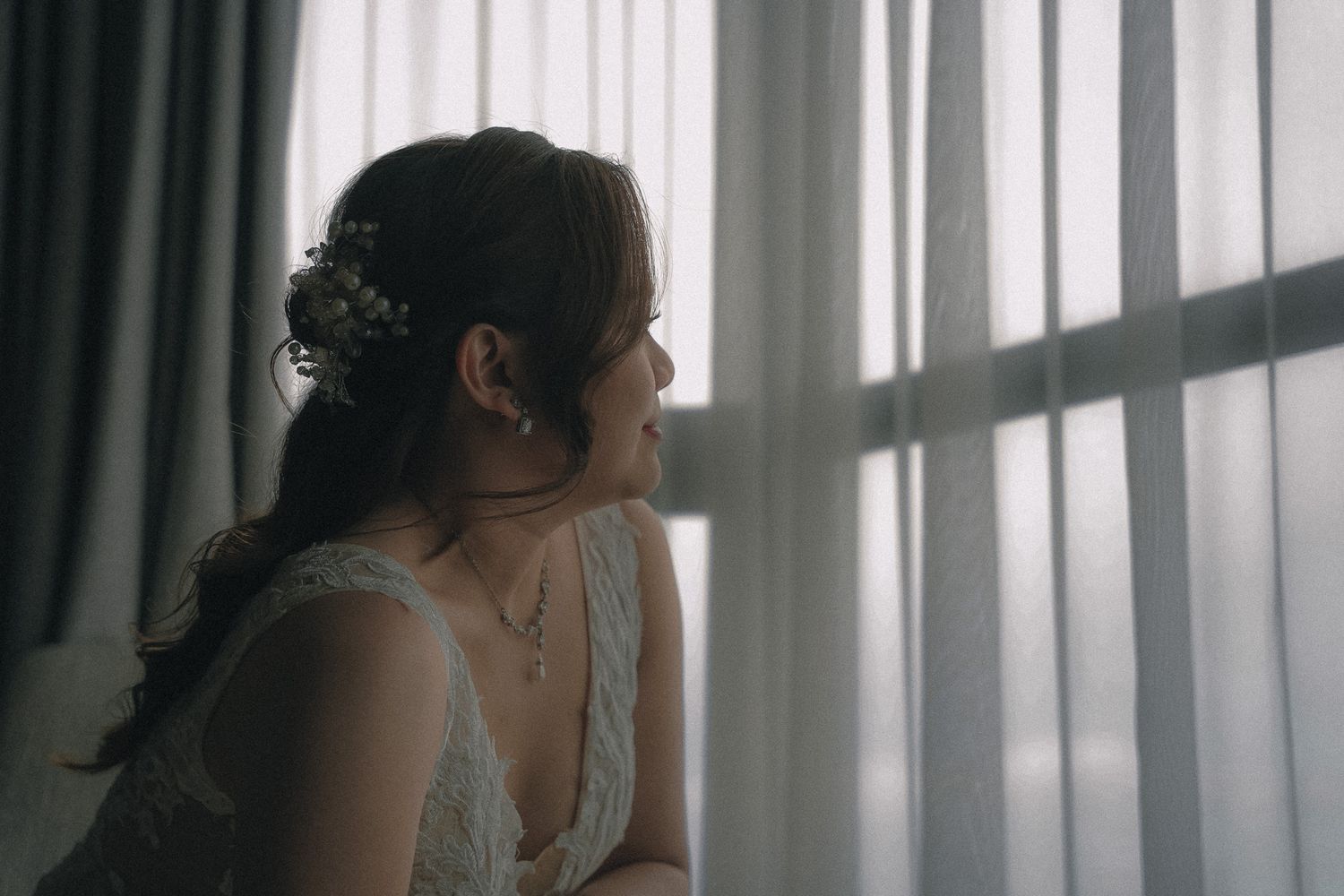 A silhouetted figure in a sleeveless dress gazes pensively through a window with sheer curtains in soft natural light.