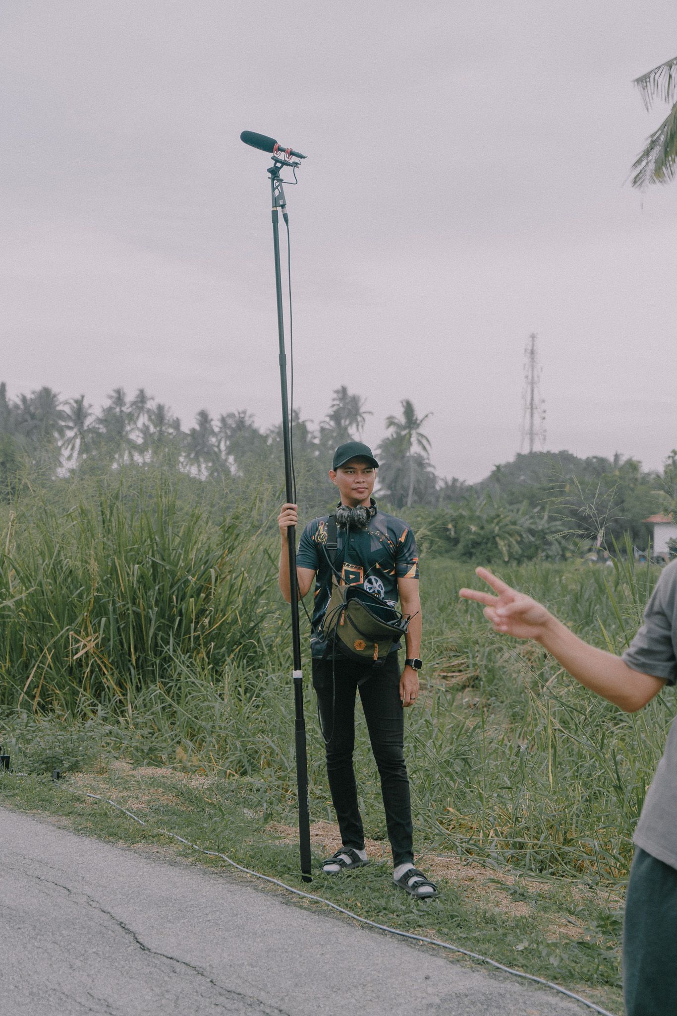 Sound technician with boom microphone pole stands on rural road with palm trees and grass in background.