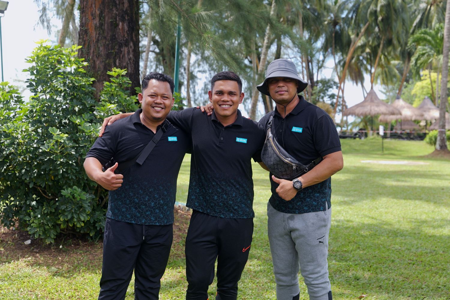 Three men in matching black uniforms smiling together in a tropical garden setting with palm trees.