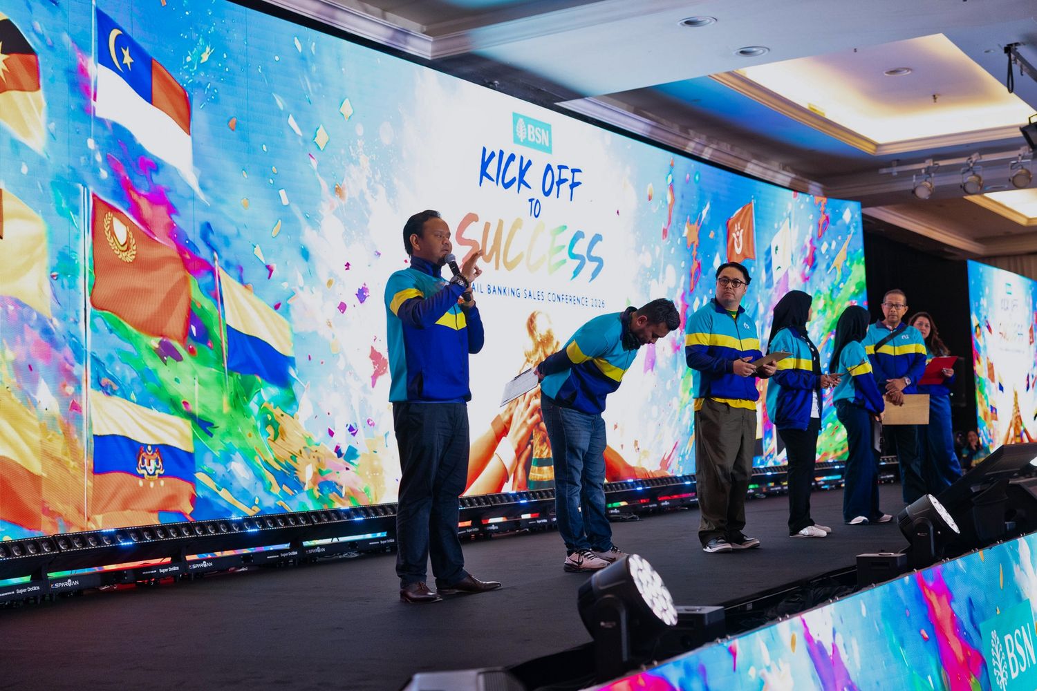 Group of people on stage at a colorful Kick Off Success event with international flags displayed on LED screen backdrop.