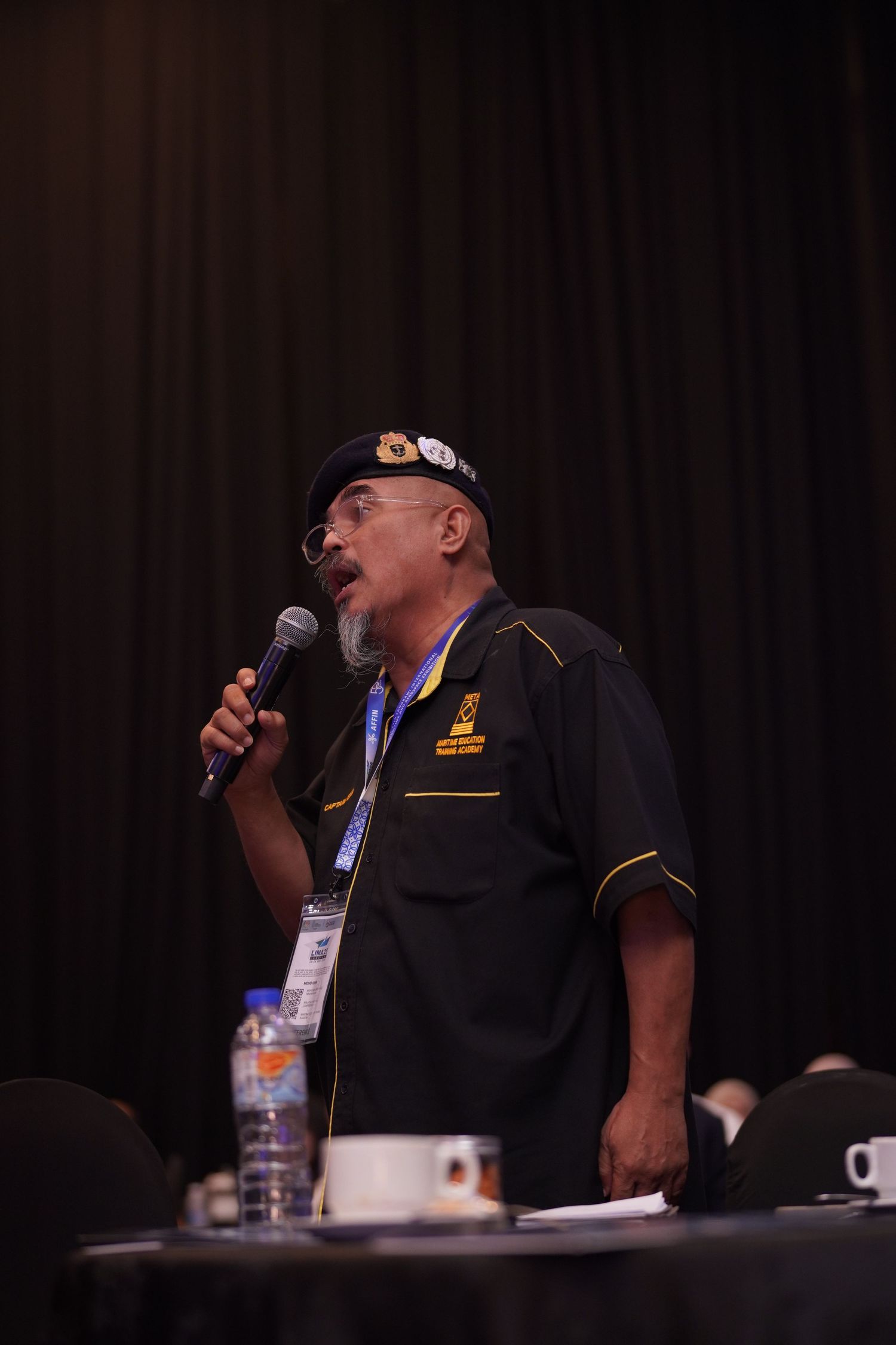 Speaker in black uniform addressing audience with microphone at conference session.