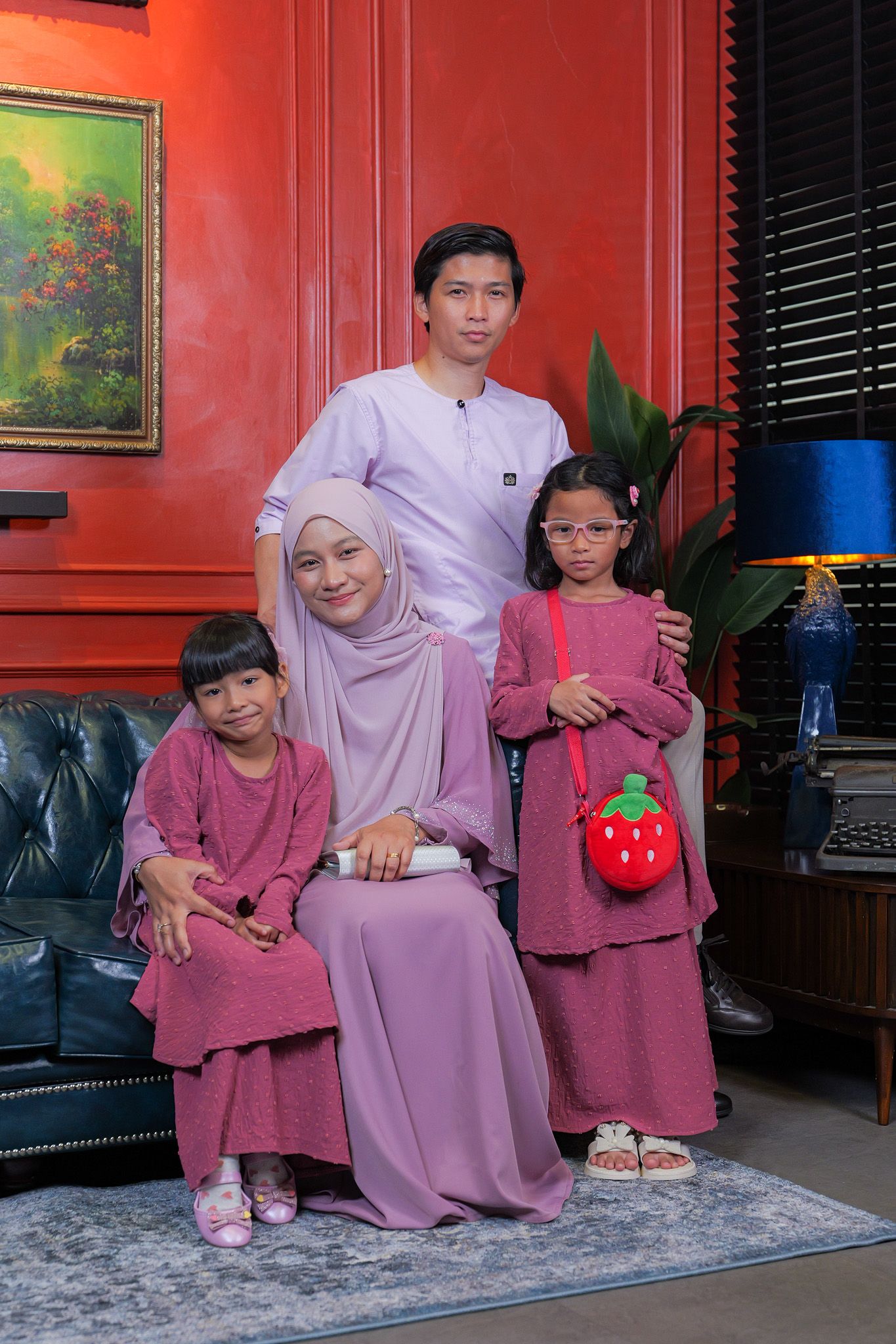 Family wearing coordinated pink and purple outfits poses together on leather couch against vibrant red wall.