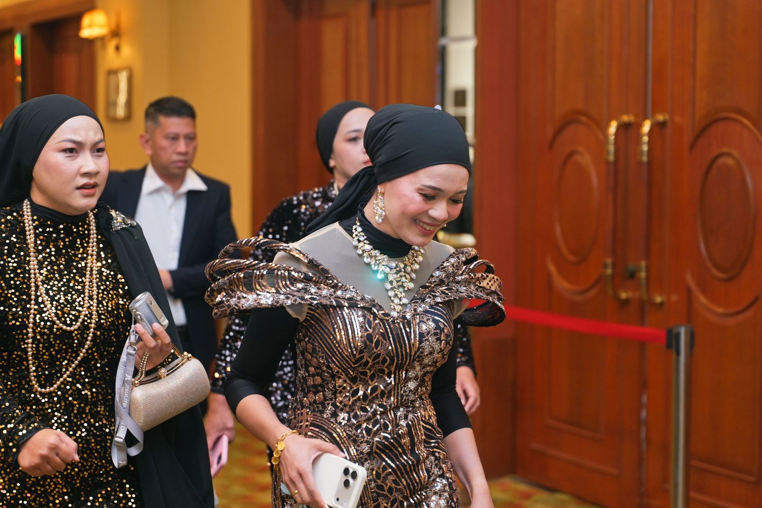 Elegantly dressed women in sequined outfits walking through a grand hotel corridor at a gala.
