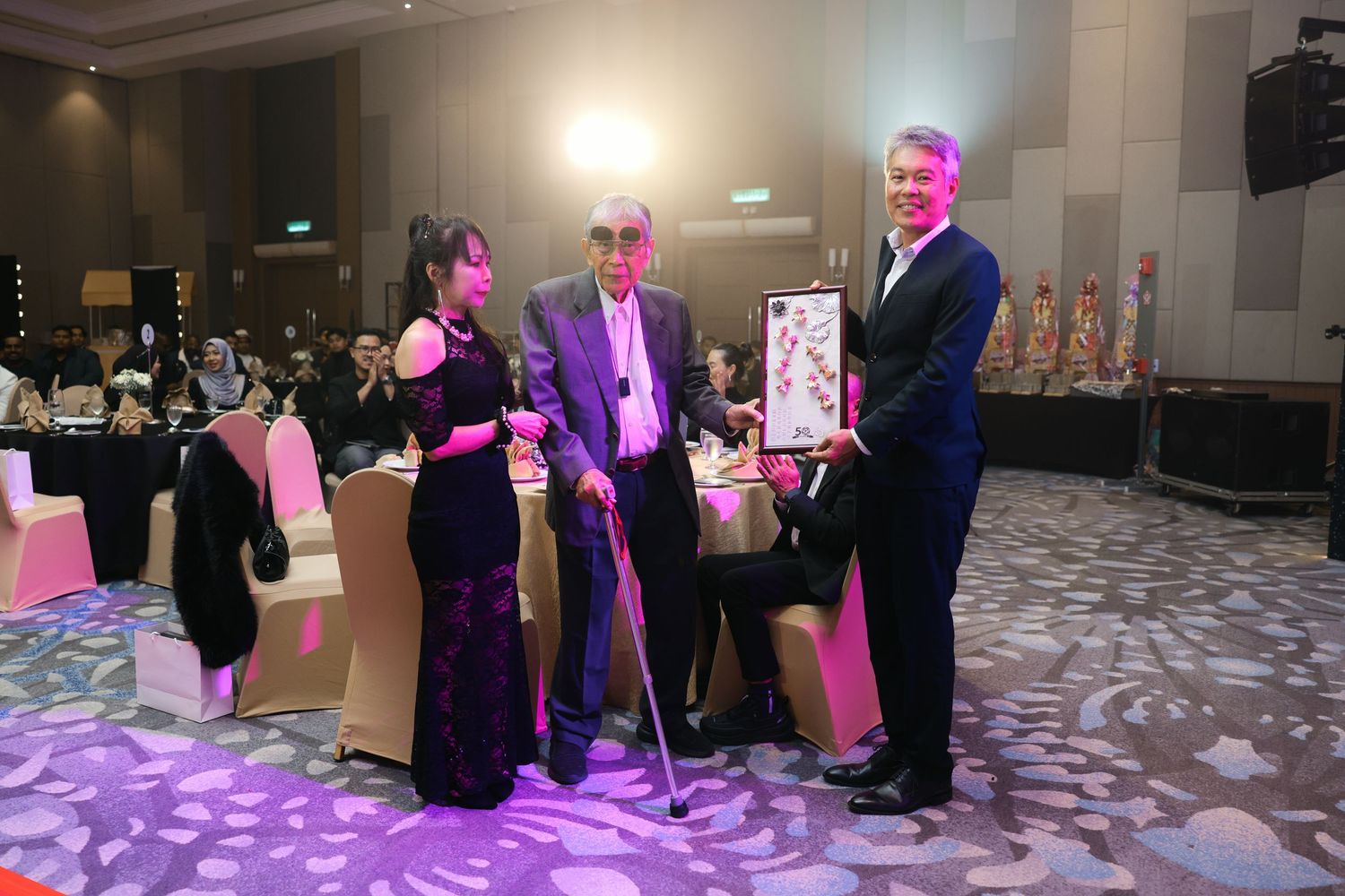 Three people presenting a framed artwork gift to an elderly man with a cane at a formal gala.
