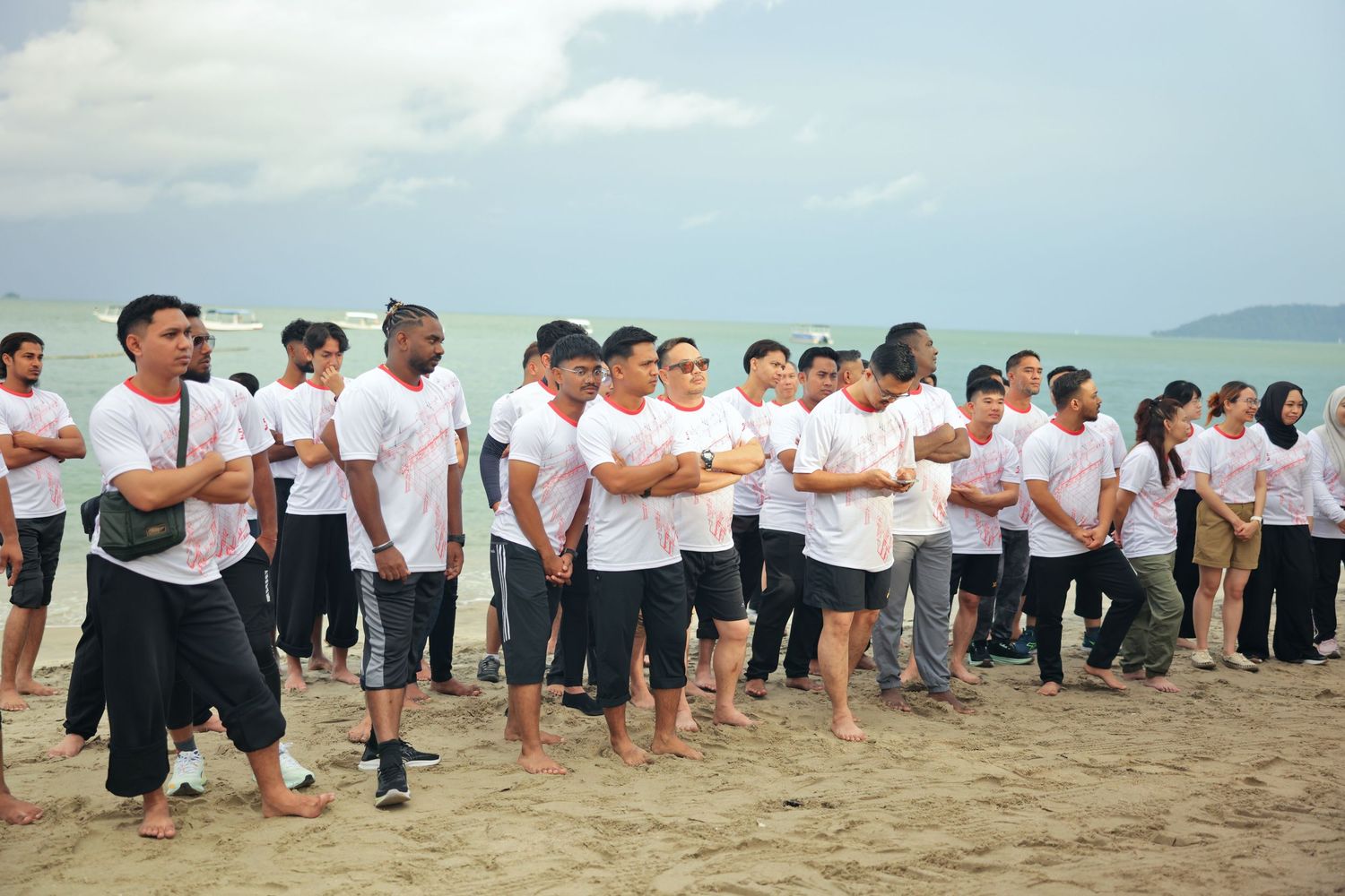 Large group of people in white shirts standing barefoot on a sandy beach during an outdoor team event.