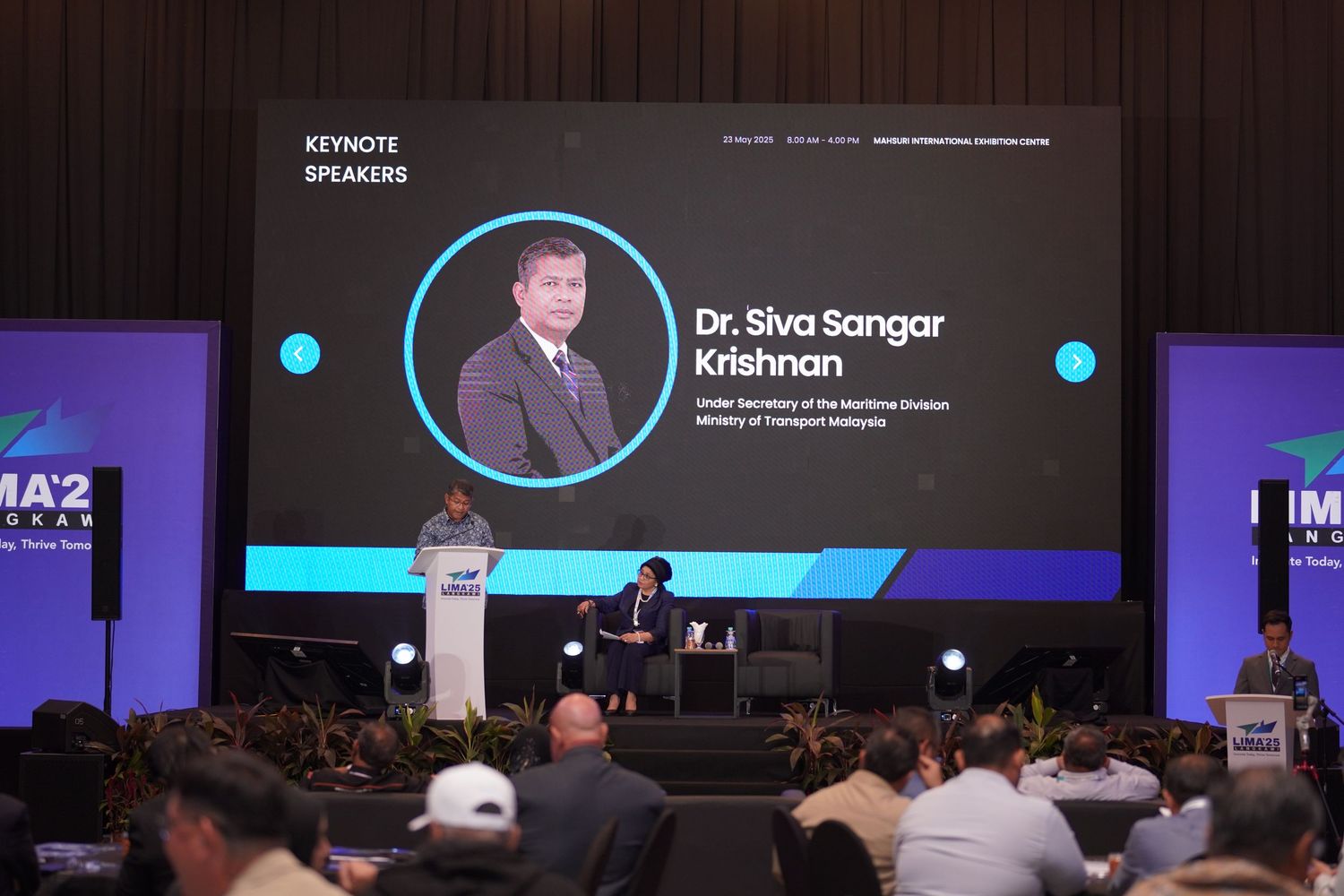 Large conference hall with speaker presentation displayed on stage screen.