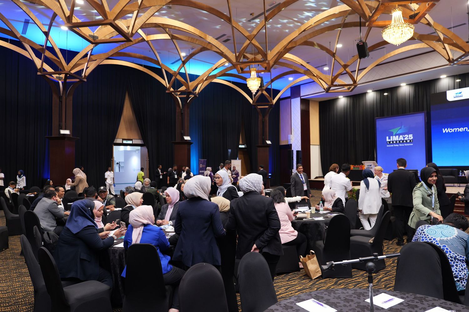 Conference attendees seated at round tables in a room with elegant wooden ceiling beams and lighting.