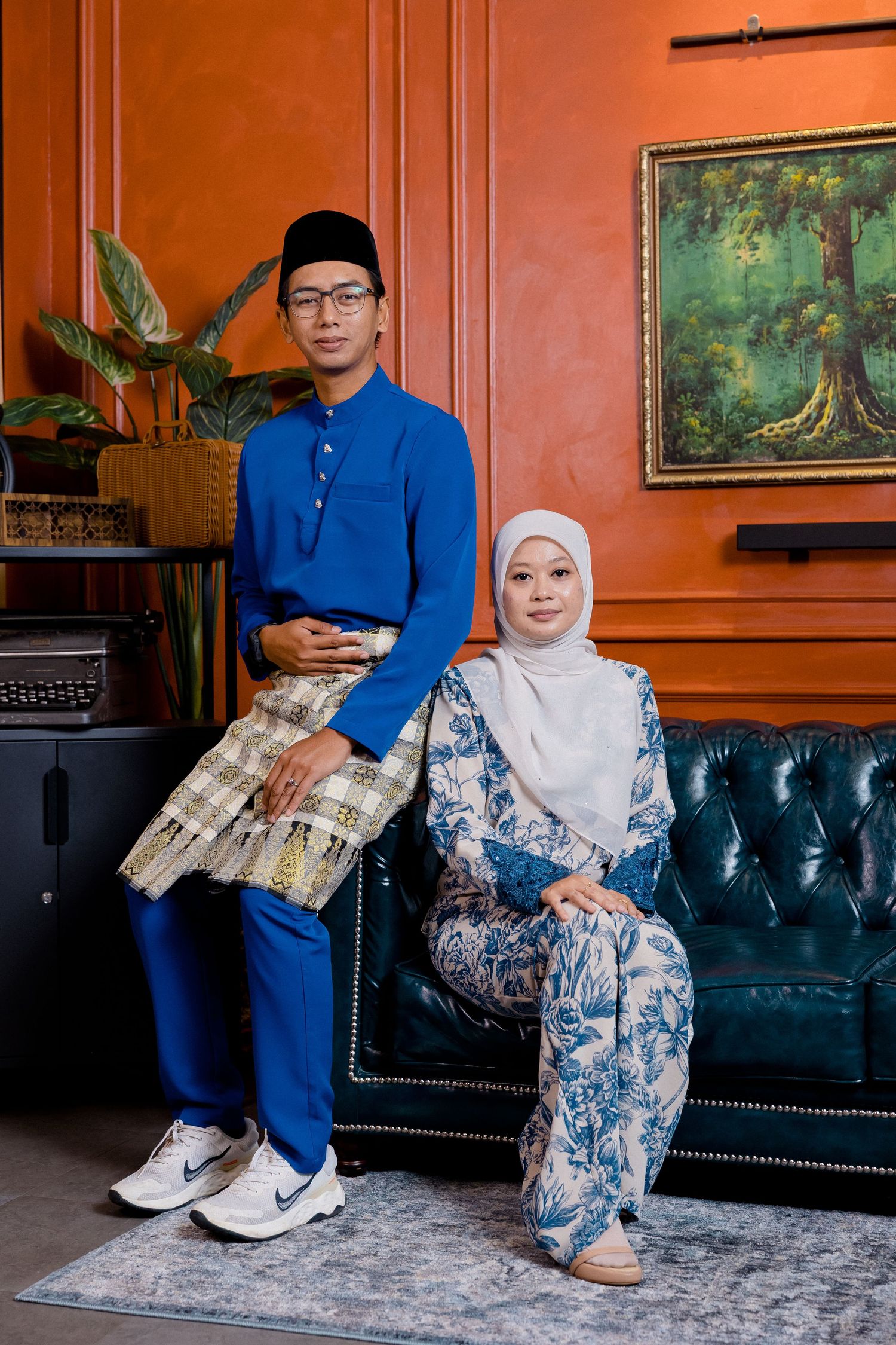 A couple sits on a tufted leather sofa in a room with orange walls and landscape paintings.
