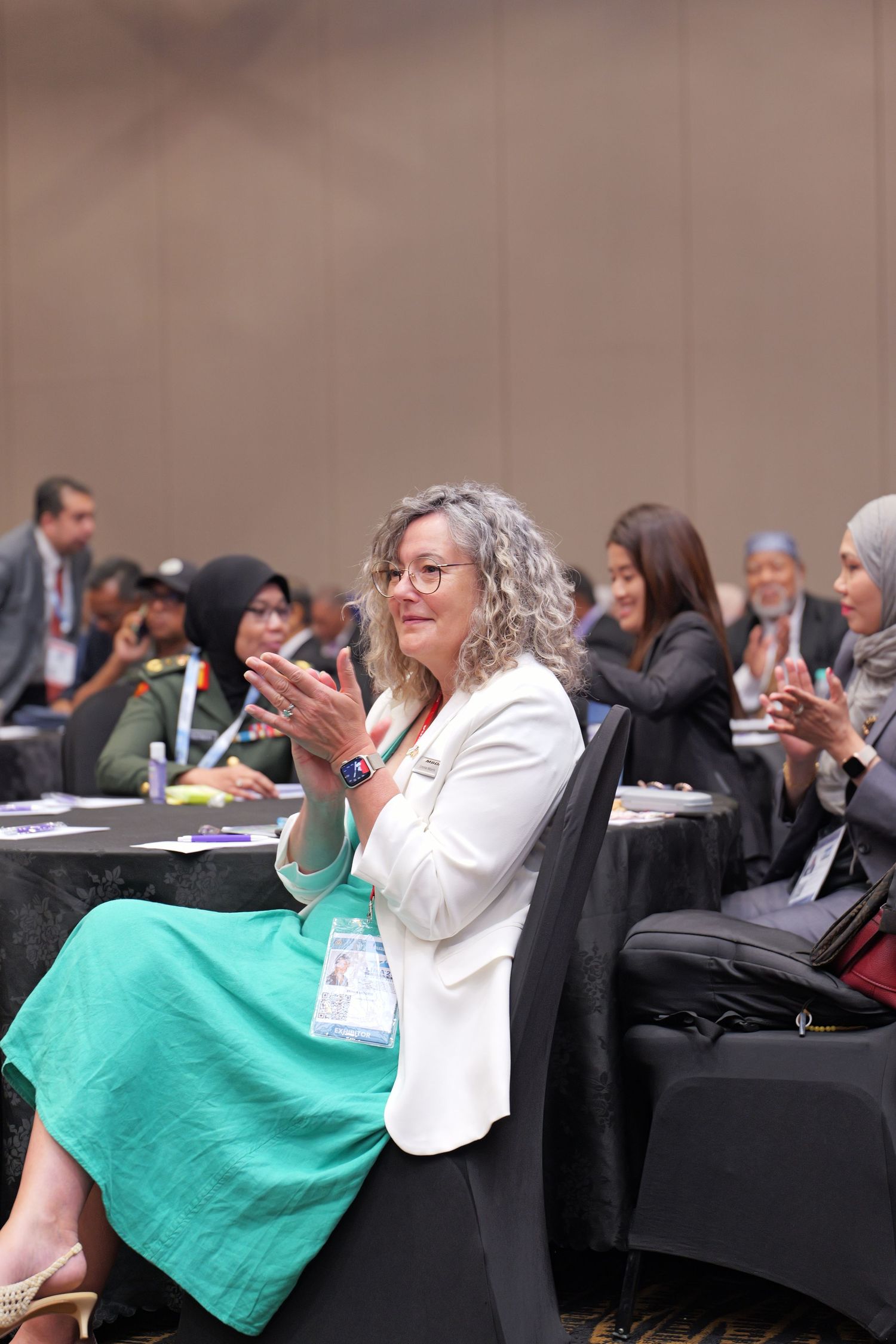 Conference attendee in white blazer and teal skirt applauds during professional business meeting.