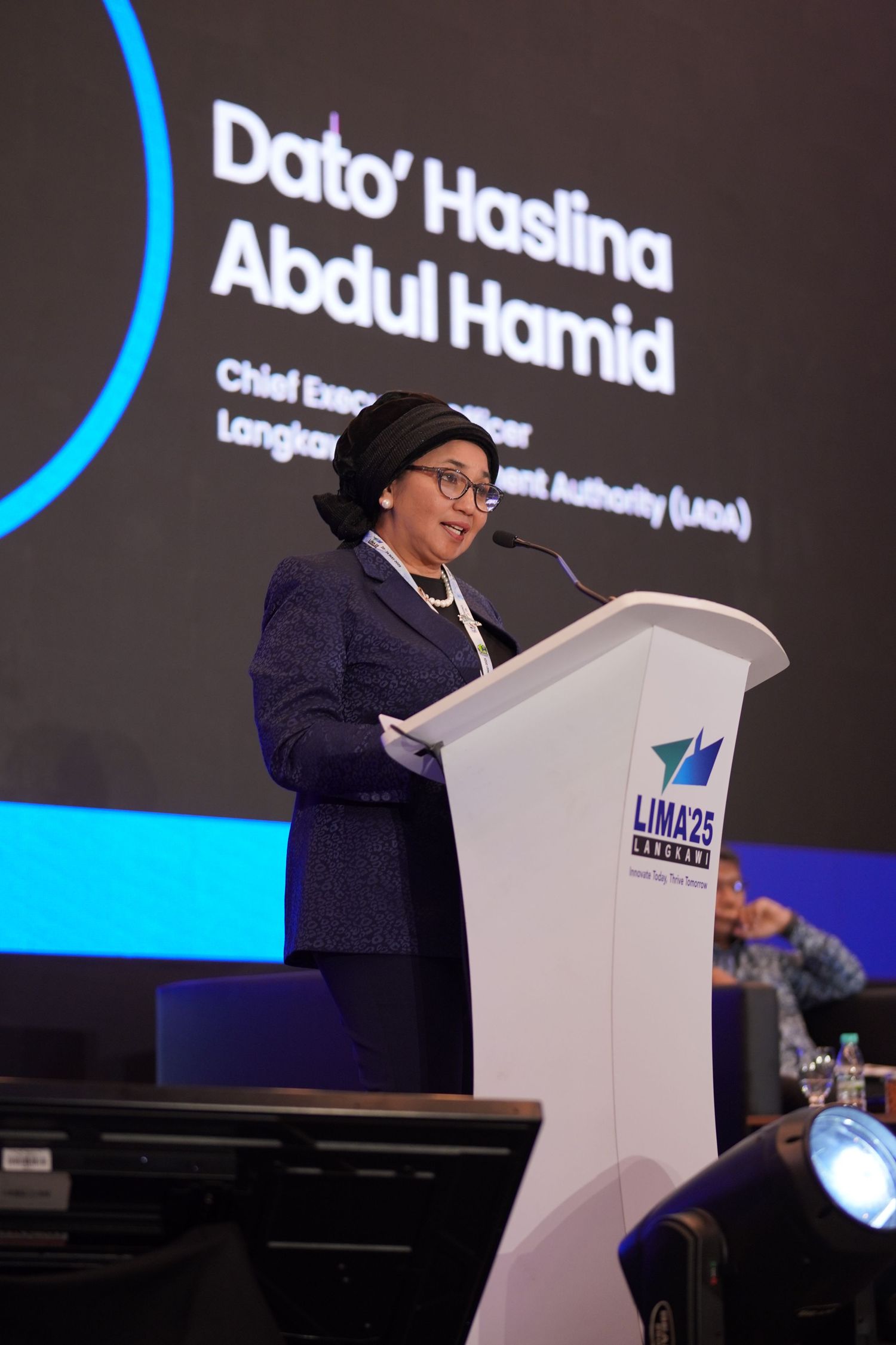 Speaker presents from a white podium with corporate branding during a professional conference.