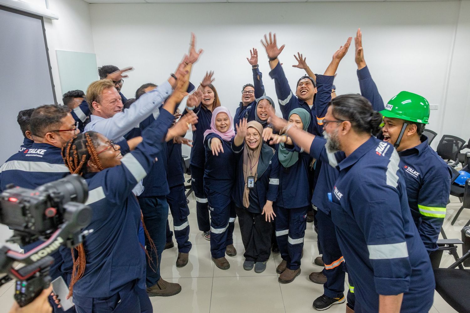 A group of workers in navy uniforms and safety gear celebrating together with raised hands in an indoor facility.