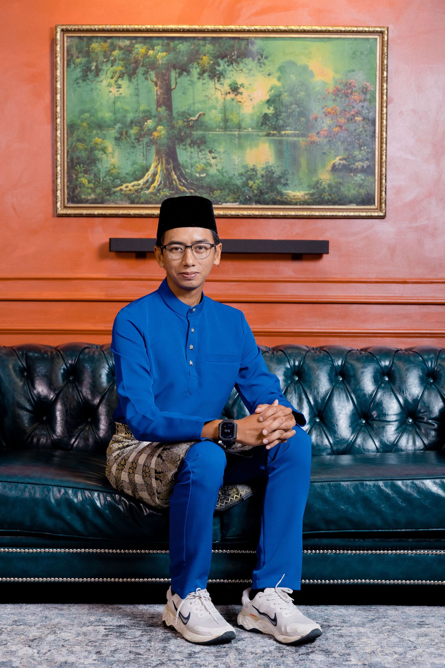 A person in a blue traditional outfit and glasses sits on a dark leather couch against an orange wall with artwork.