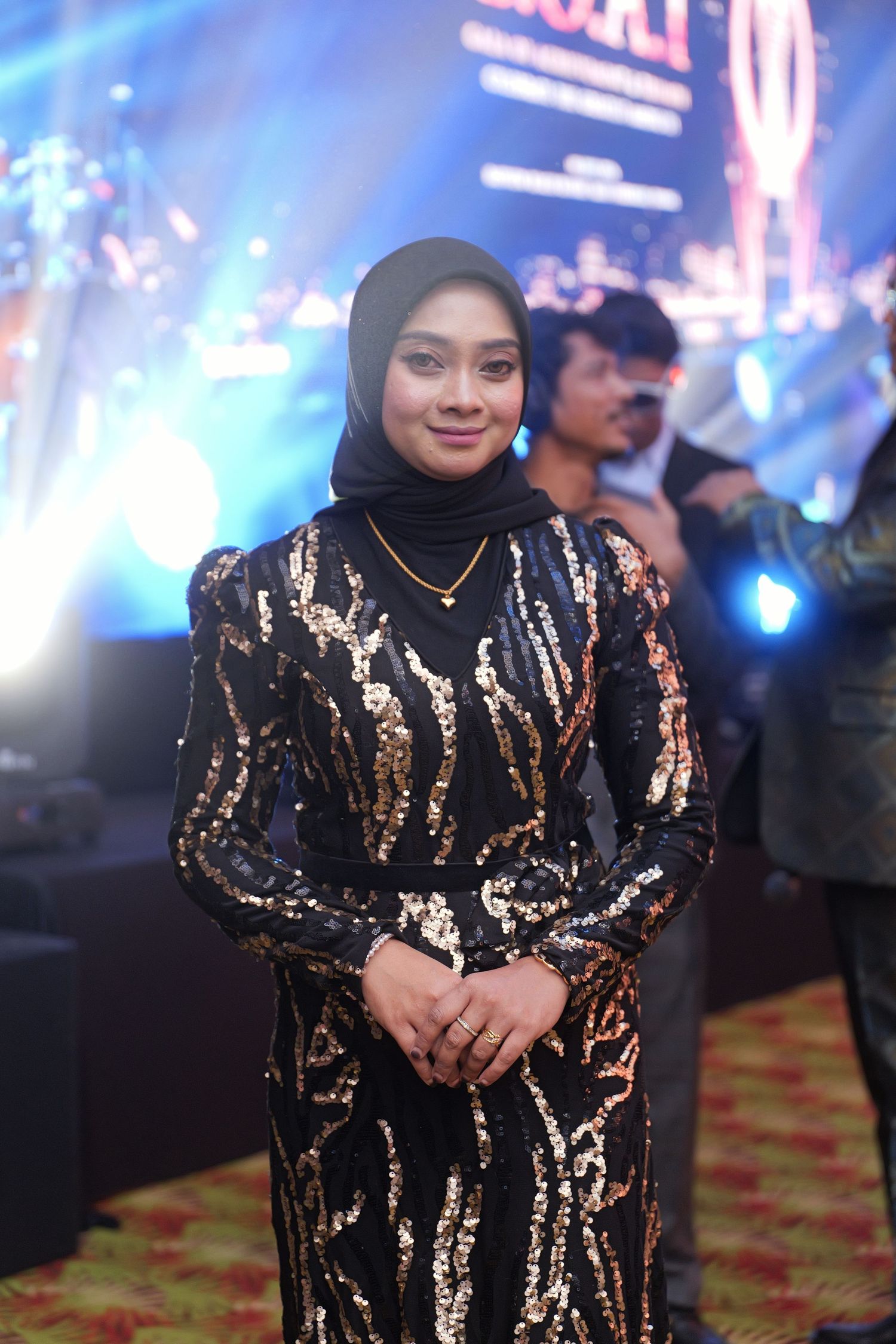 Woman in black and gold sequin dress with hijab smiling at formal awards event.